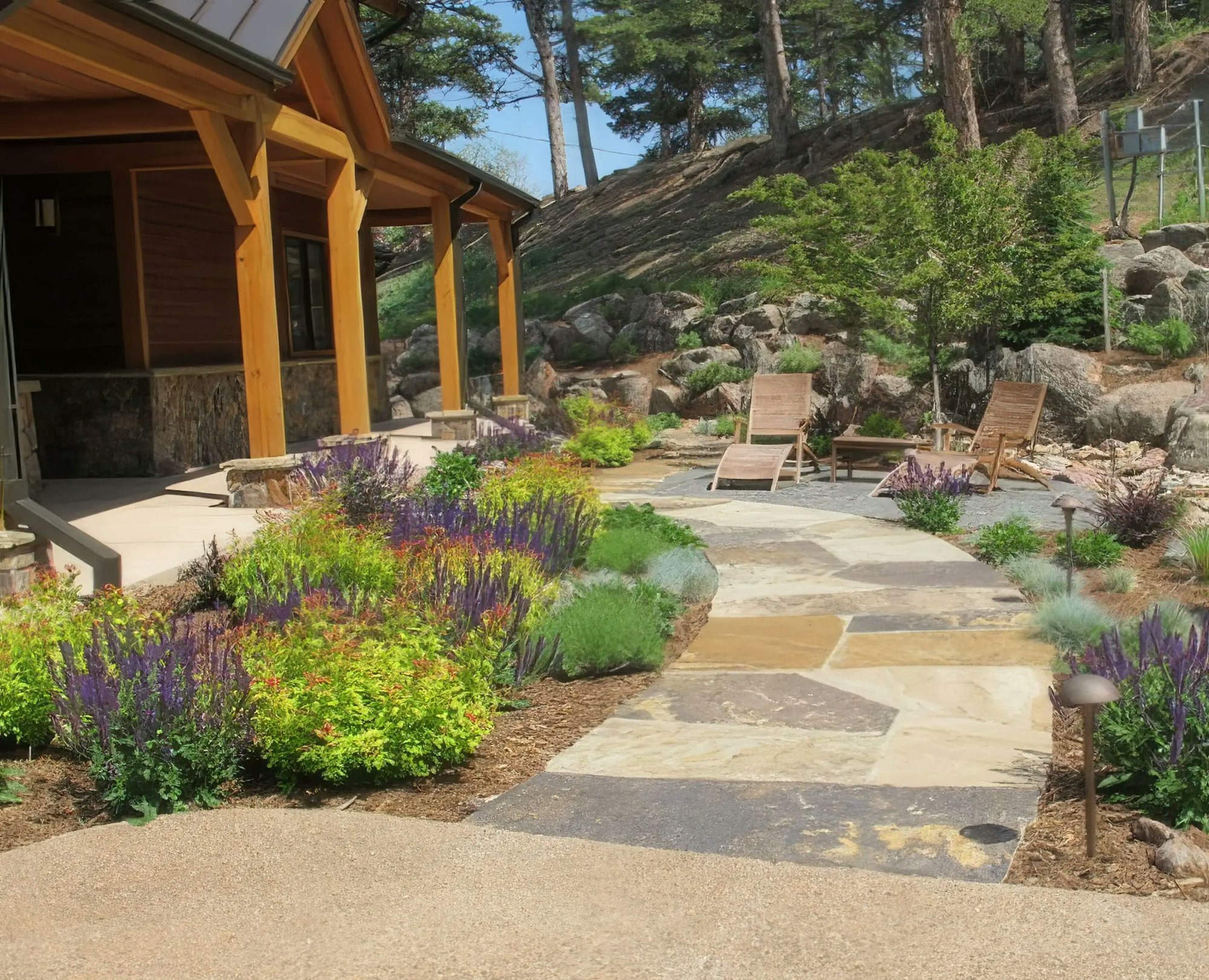 A landscaped yard with colorful flowers, a stone pathway, and wooden lounge chairs near a modern home. Trees and rocks in the background.