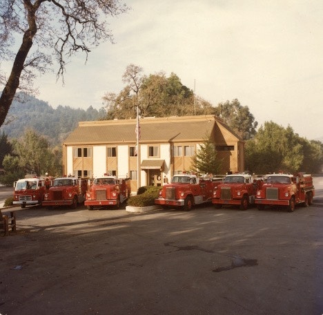 A fire station with several red fire trucks parked outside, surrounded by trees and a clear sky.