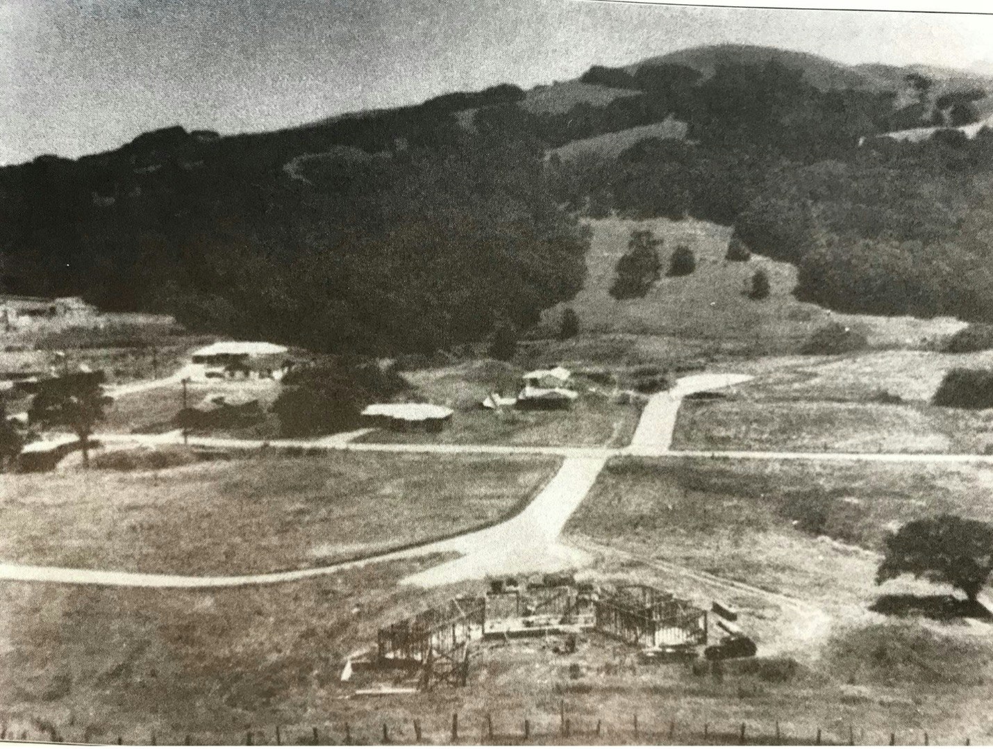 A black-and-white image showing a rural landscape with roads, houses, and a partially constructed structure in a grassy area.