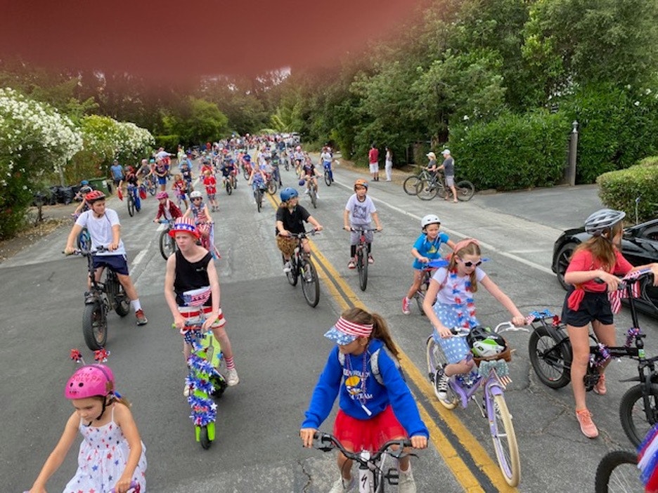 A vibrant group of kids riding bicycles in a festive parade, many wearing patriotic decorations and enjoying the outdoors.