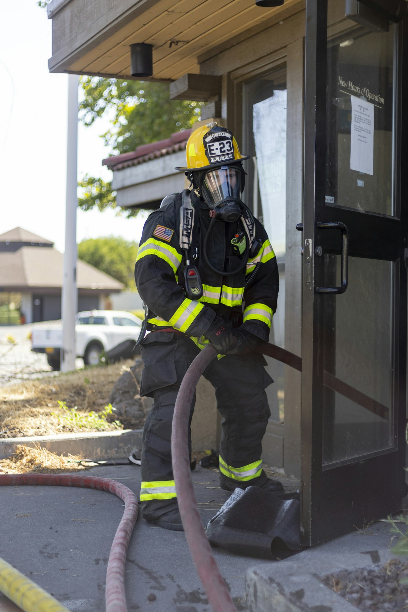 A firefighter in gear holds a hose outside a building, preparing for action.