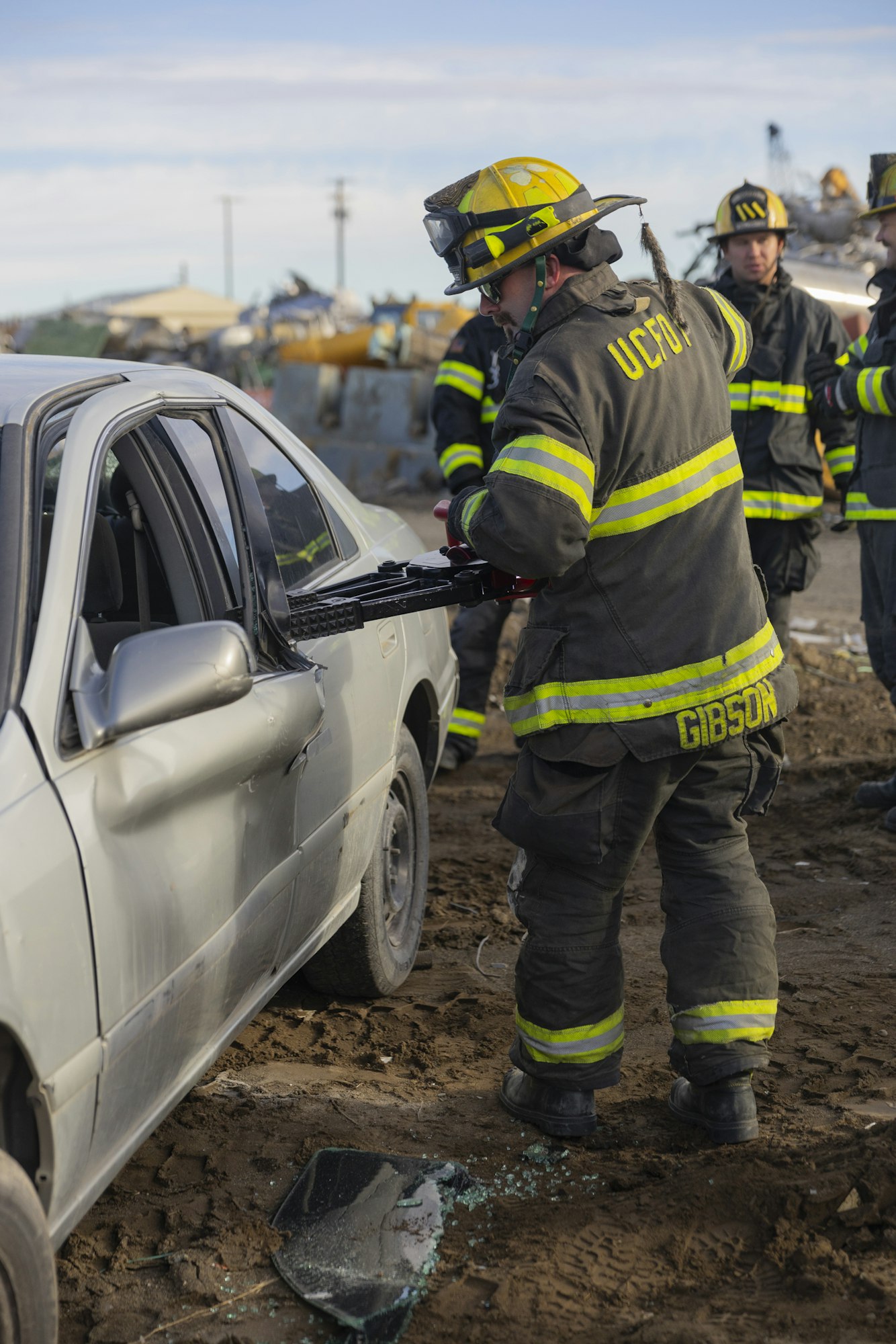 Firefighter using hydraulic tool on a car door; team in background.