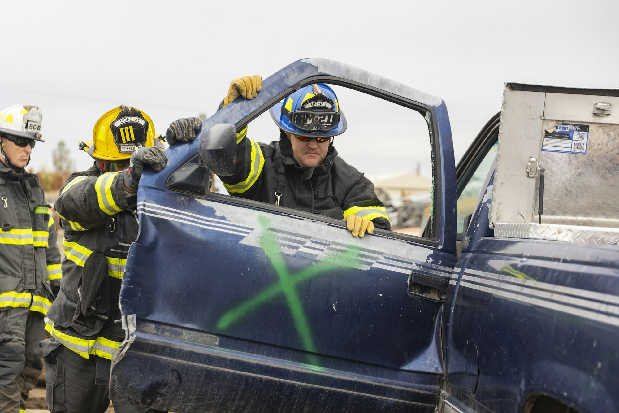 Firefighters in gear handle a blue vehicle door marked with a green "X."