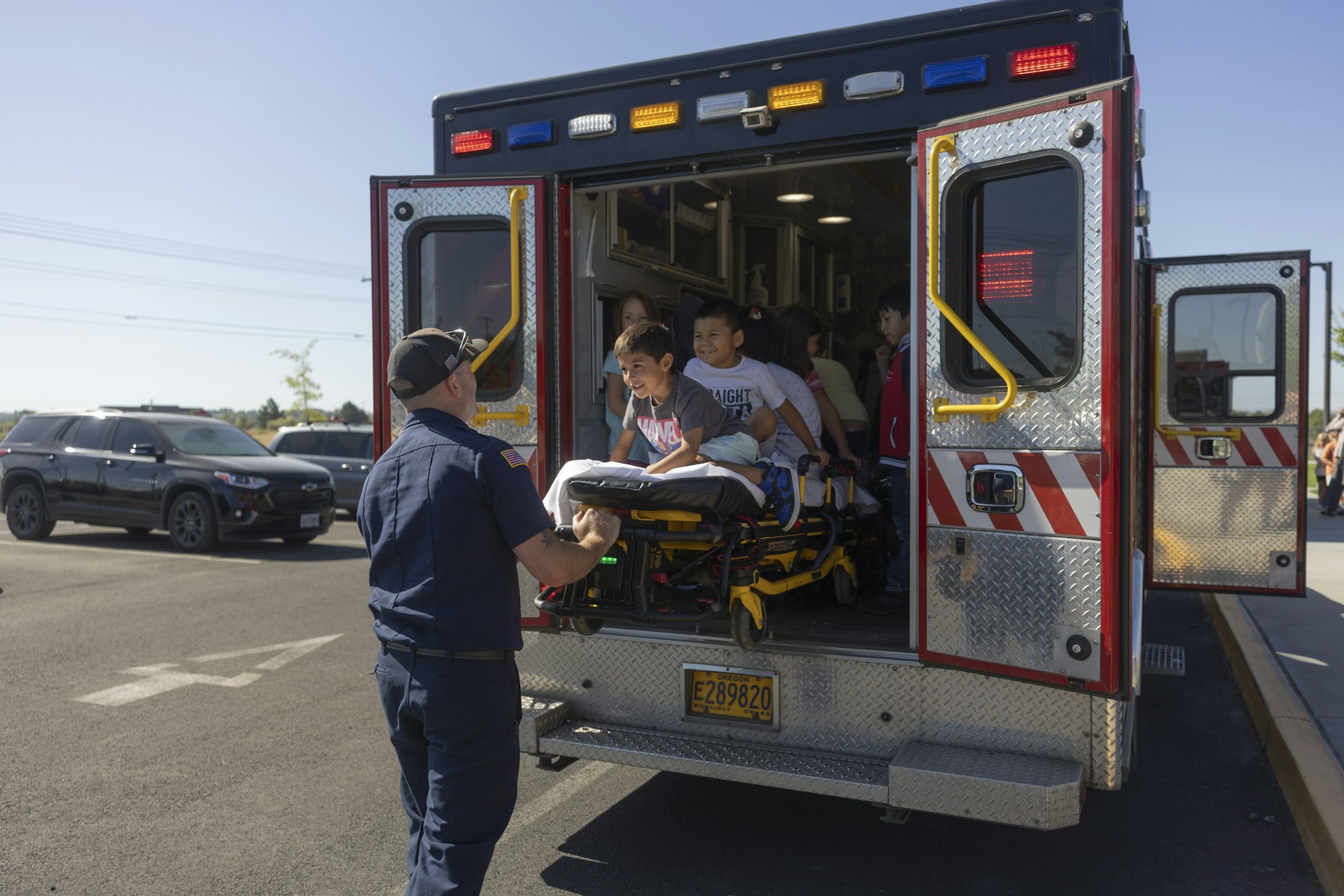 A group of children sits inside an ambulance, with one child on a stretcher, as a uniformed adult assists outside.