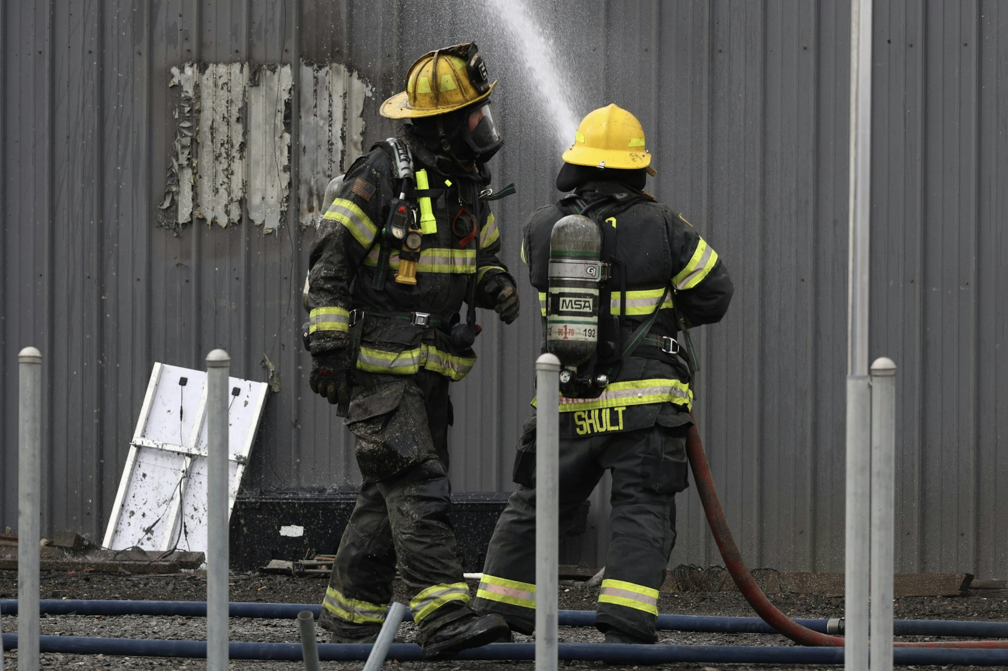 Two firefighters in gear, one spraying water, in front of a metal wall with peeling paint.