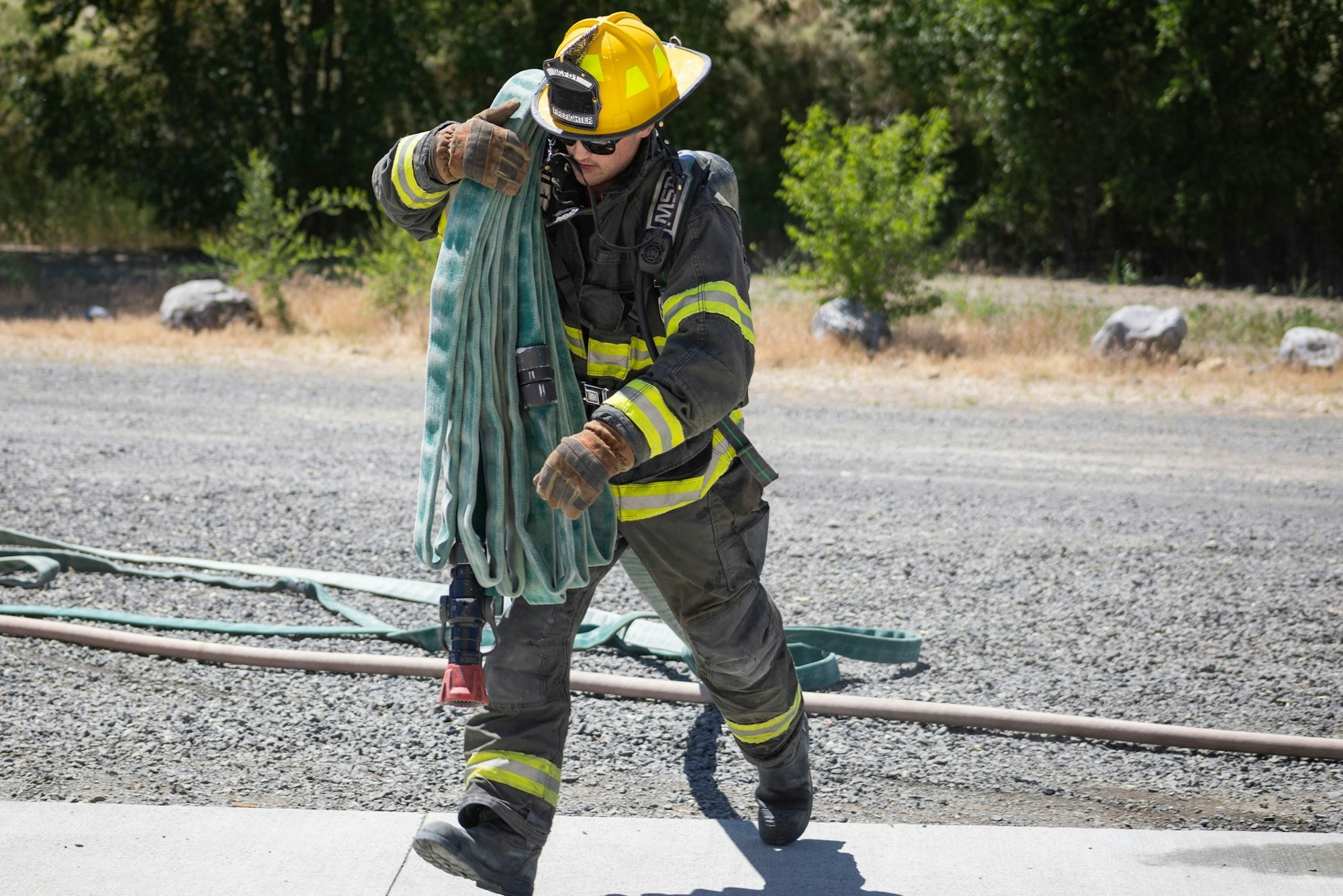A firefighter in full gear carries a coiled hose outdoors on a sunny day.