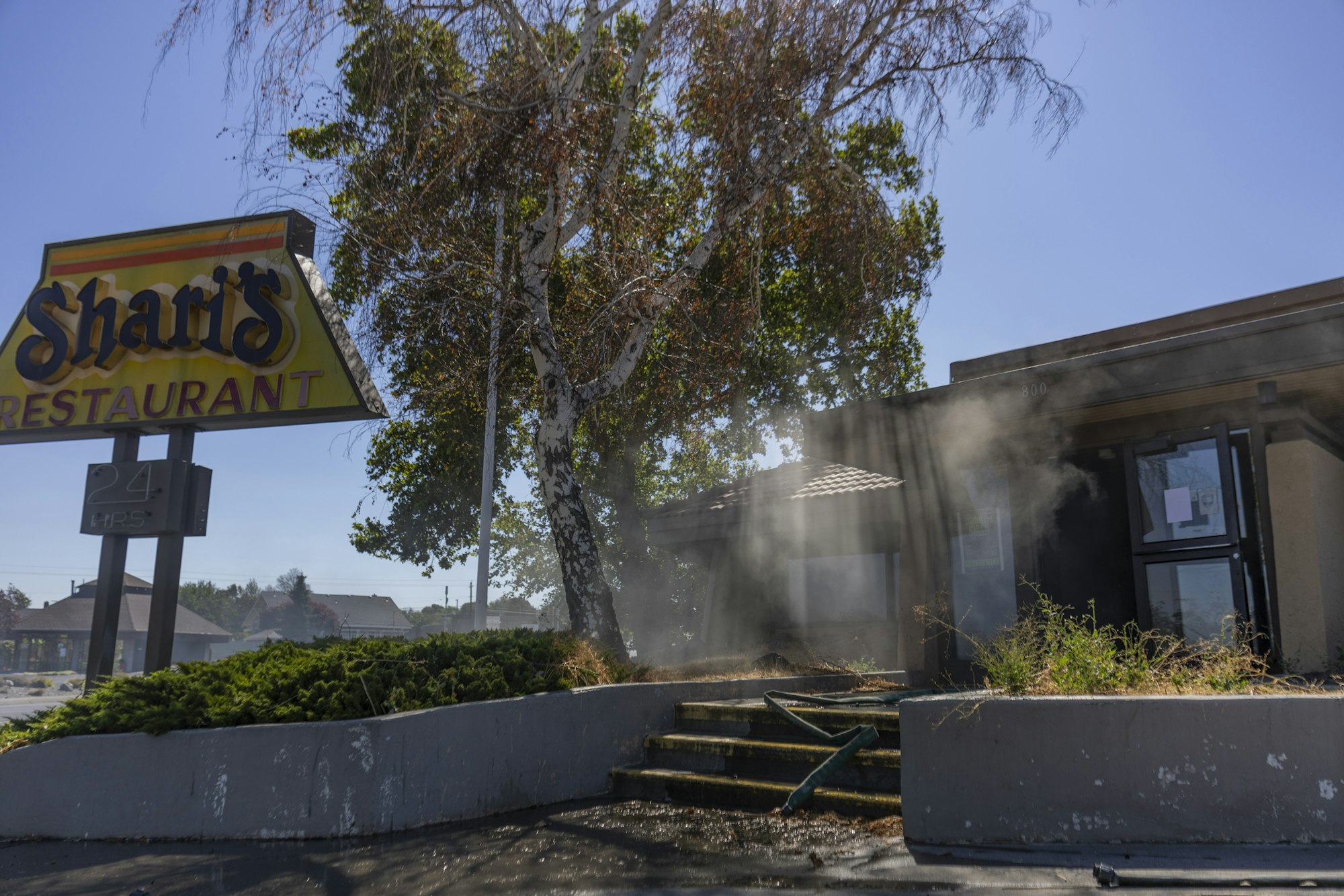 Abandoned restaurant with a "Shari's" sign, overgrown plants, and smoke or haze near the entrance under a blue sky.