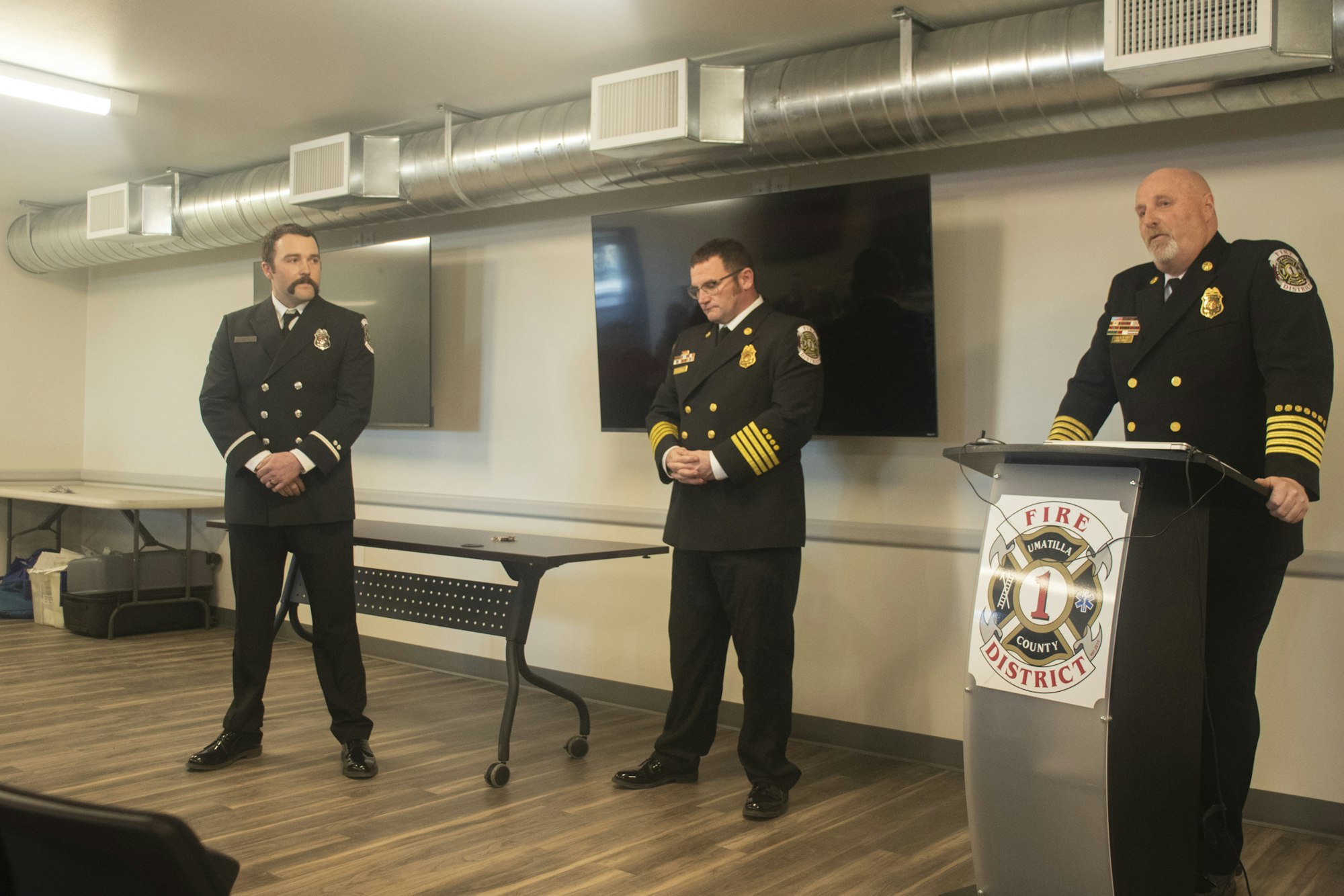 Three uniformed fire officers standing indoors, one at a podium with a "Fire District" logo.
