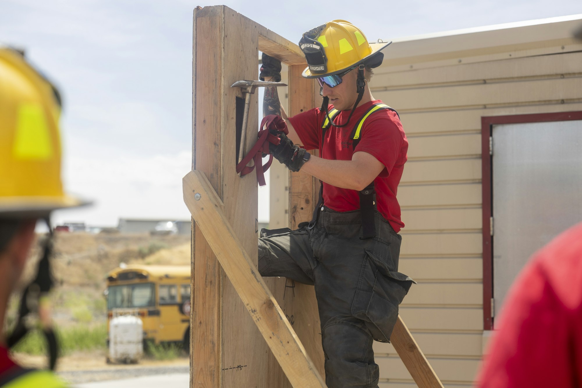 A person in a hard hat and gloves is working with wood and tools outdoors while another observes.