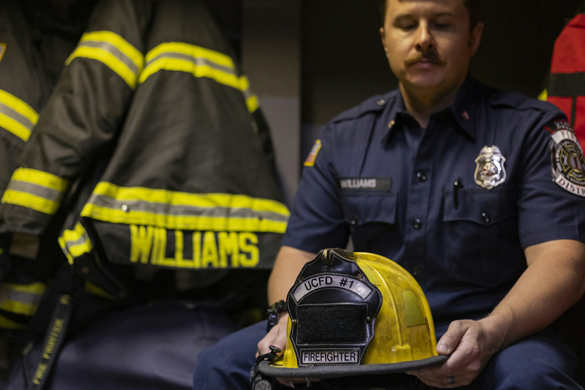 Firefighter in uniform holds a yellow helmet with "UCFD #1" label; fire jackets with "Williams" hang nearby.
