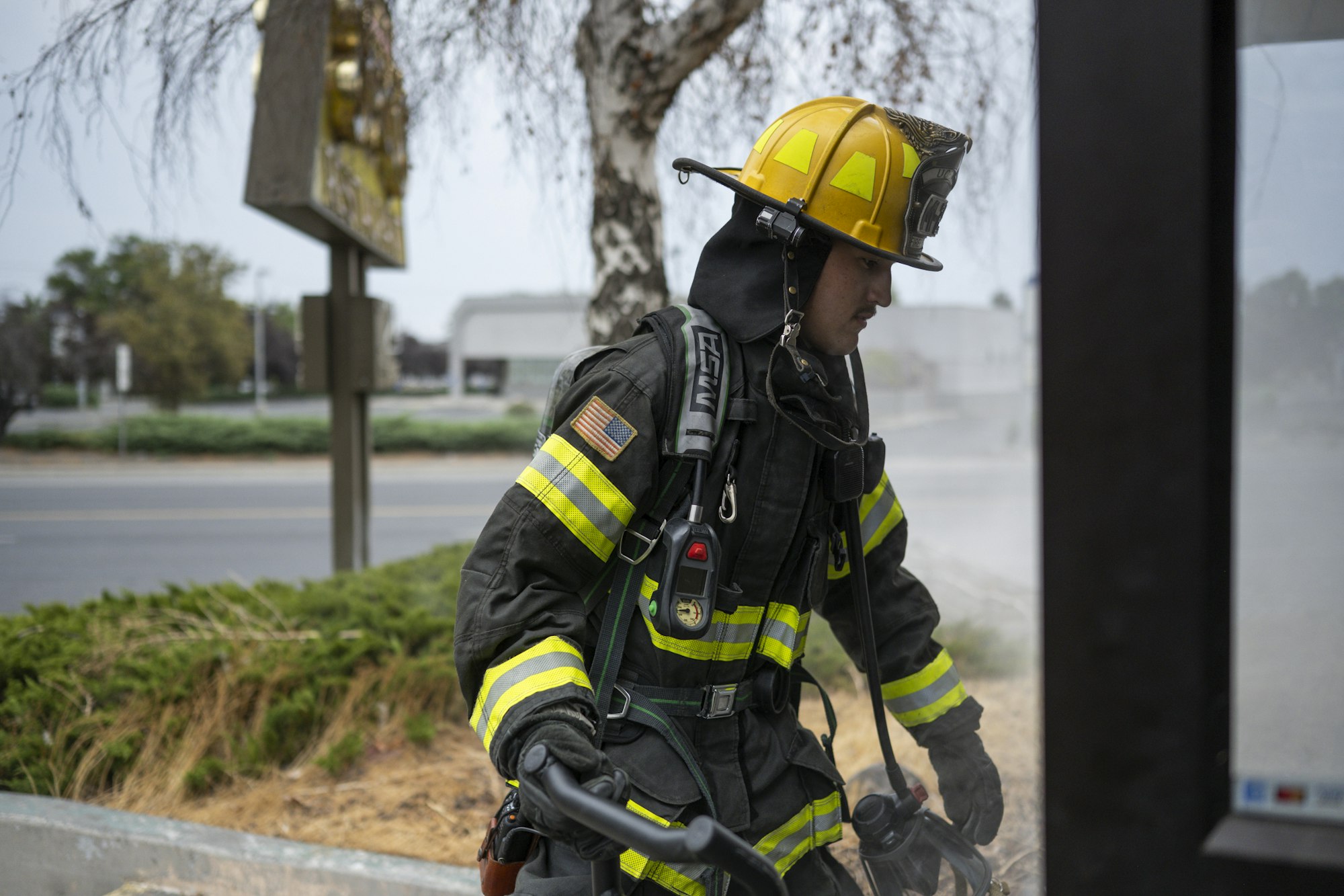 A firefighter in full gear with helmet and equipment, standing outside near a road.