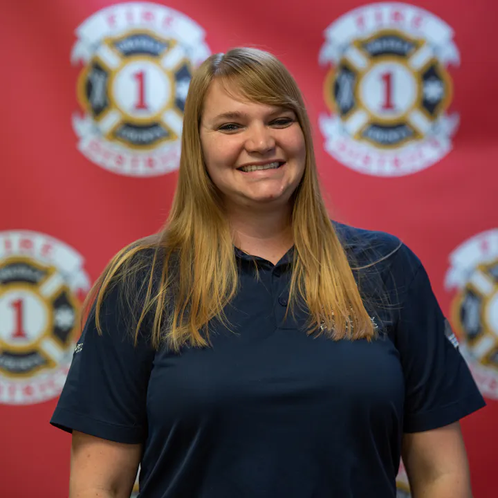 A smiling person in a dark polo shirt stands in front of a red background featuring fire department logos.