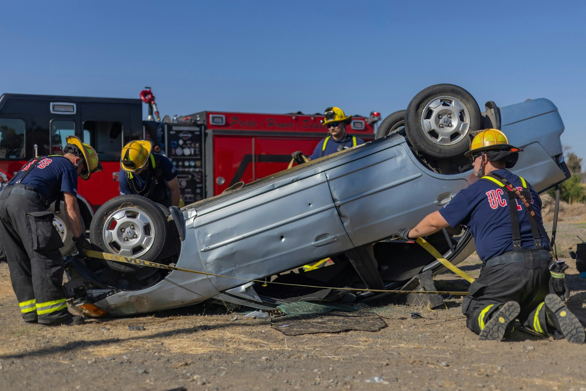 Firefighters work on an overturned car scene, using straps and tools, with a fire truck in the background.