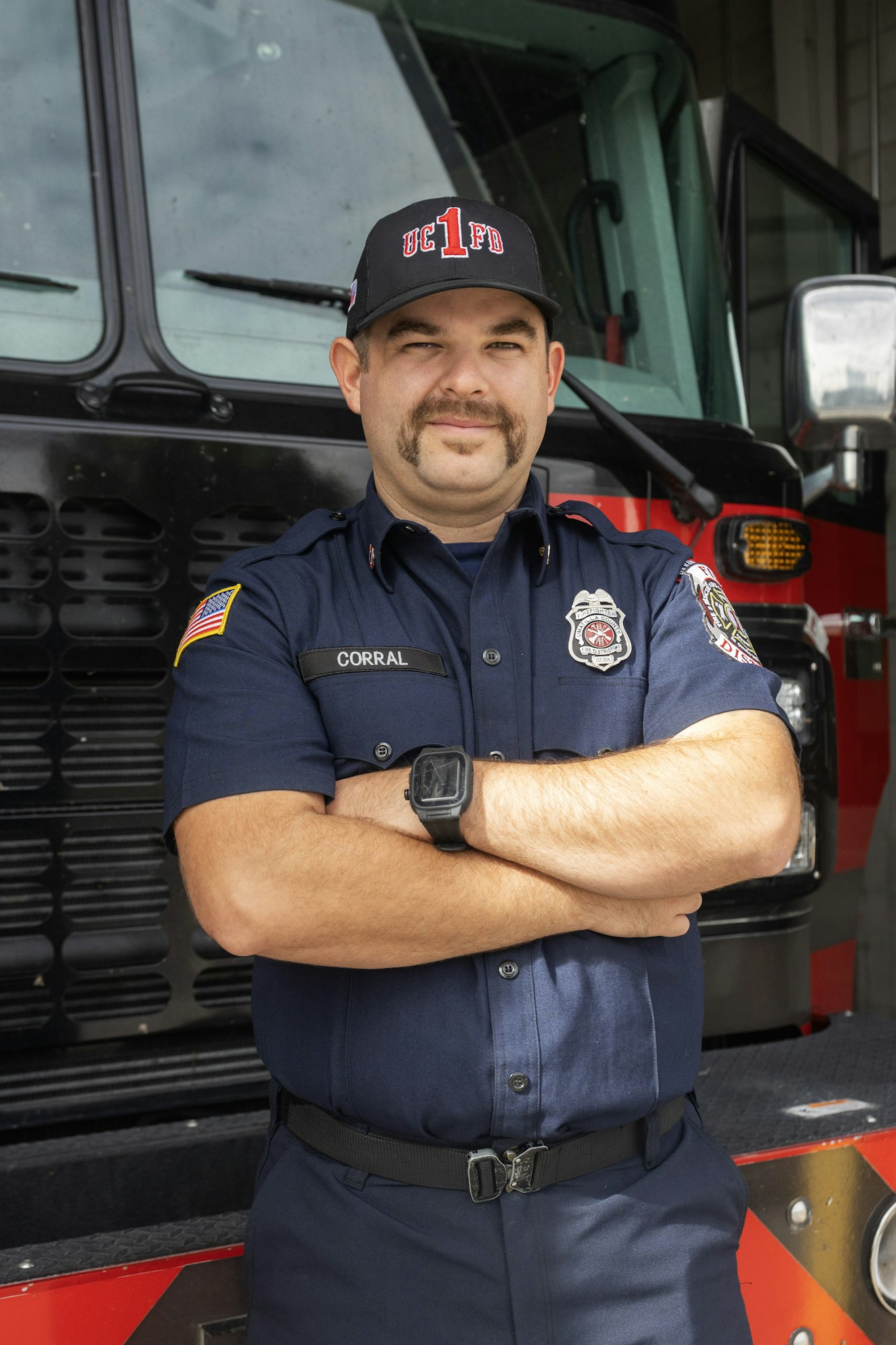 A firefighter in uniform stands with arms crossed in front of a fire truck.