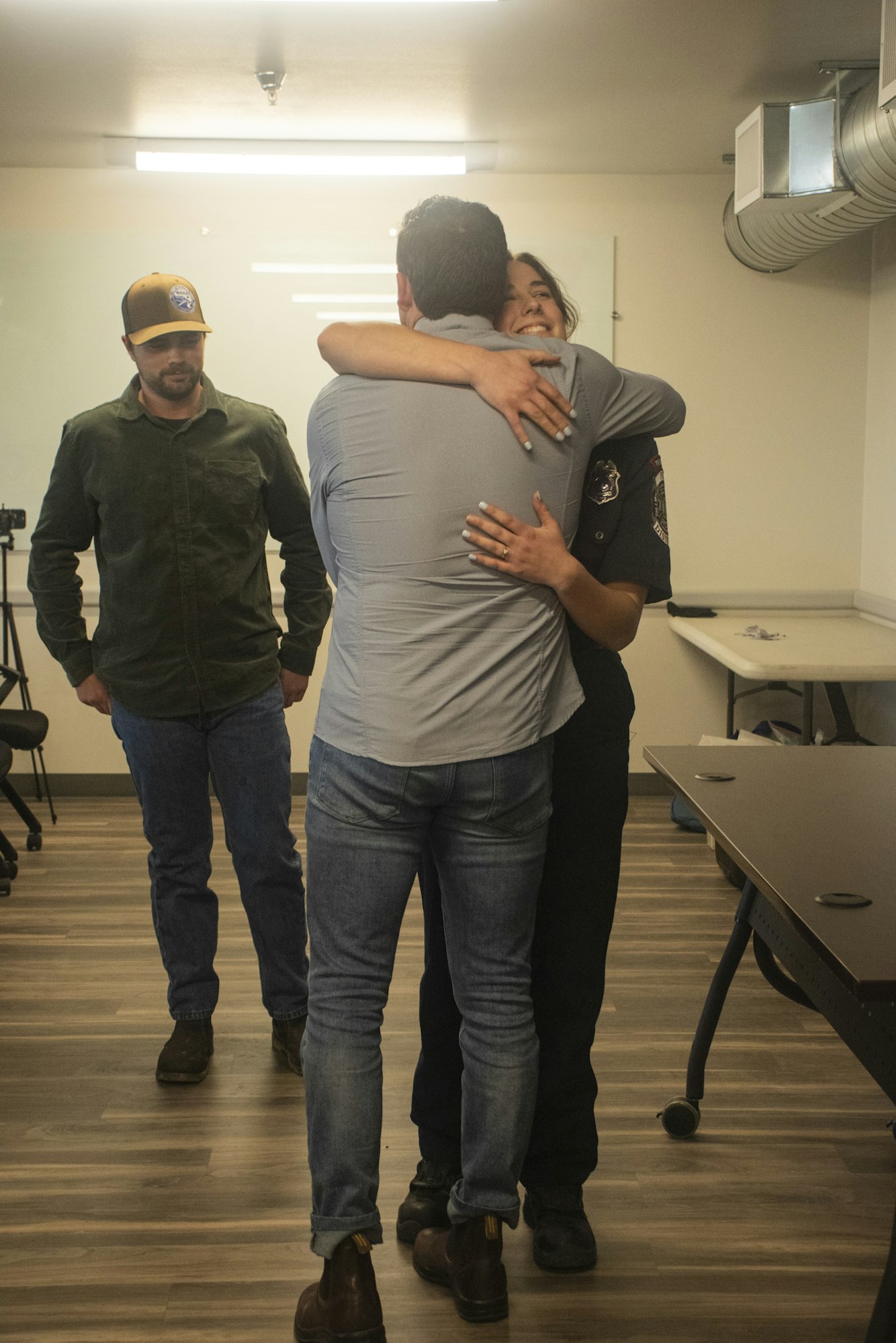 Three people in a room; one in uniform hugging another person, while the third looks on.