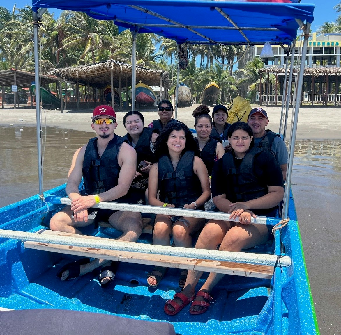 A group of people wearing life jackets sitting on a boat near a beach with palm trees in the background.