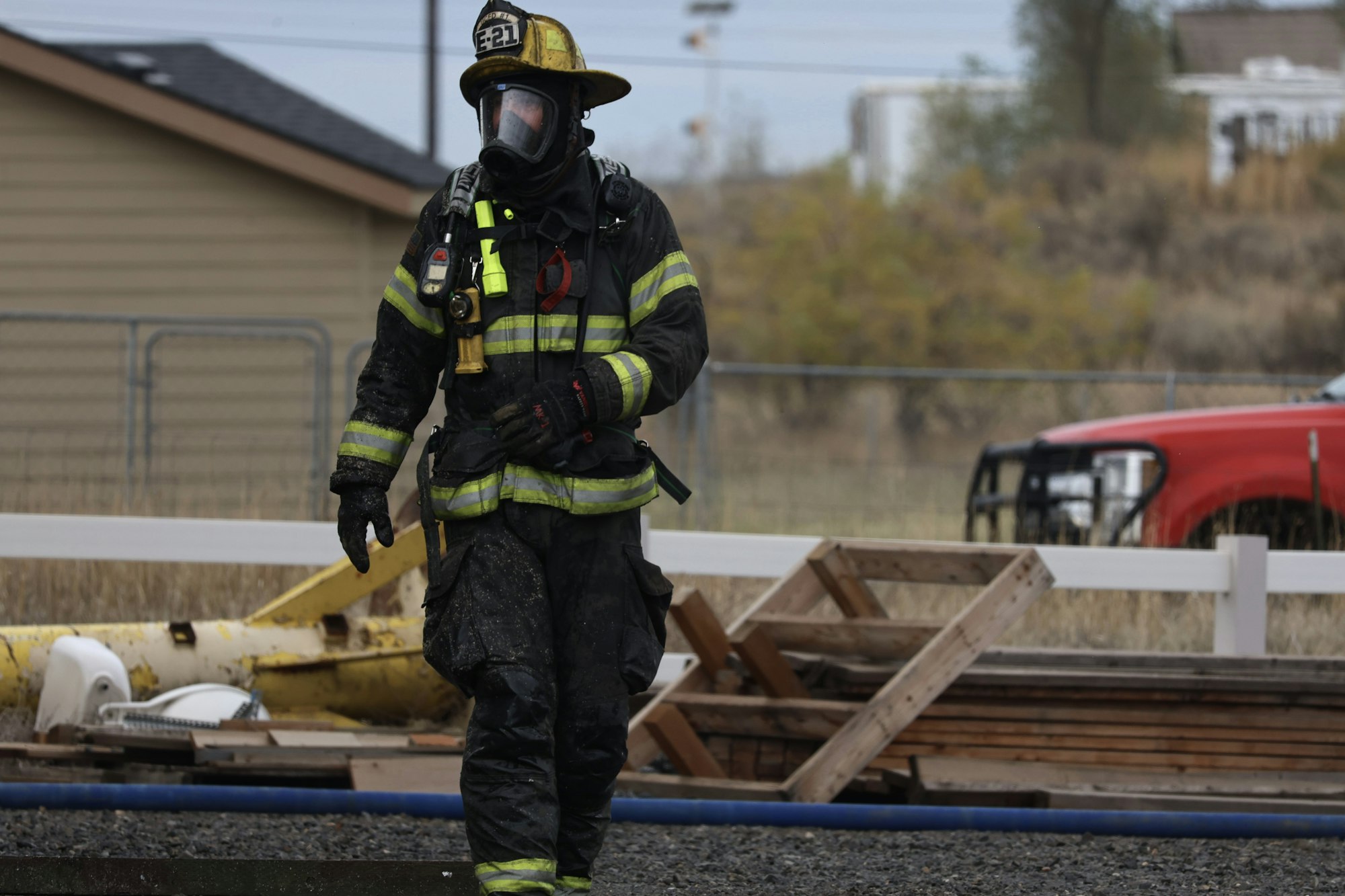 A firefighter in full gear, including helmet and mask, walks near debris and a red vehicle.