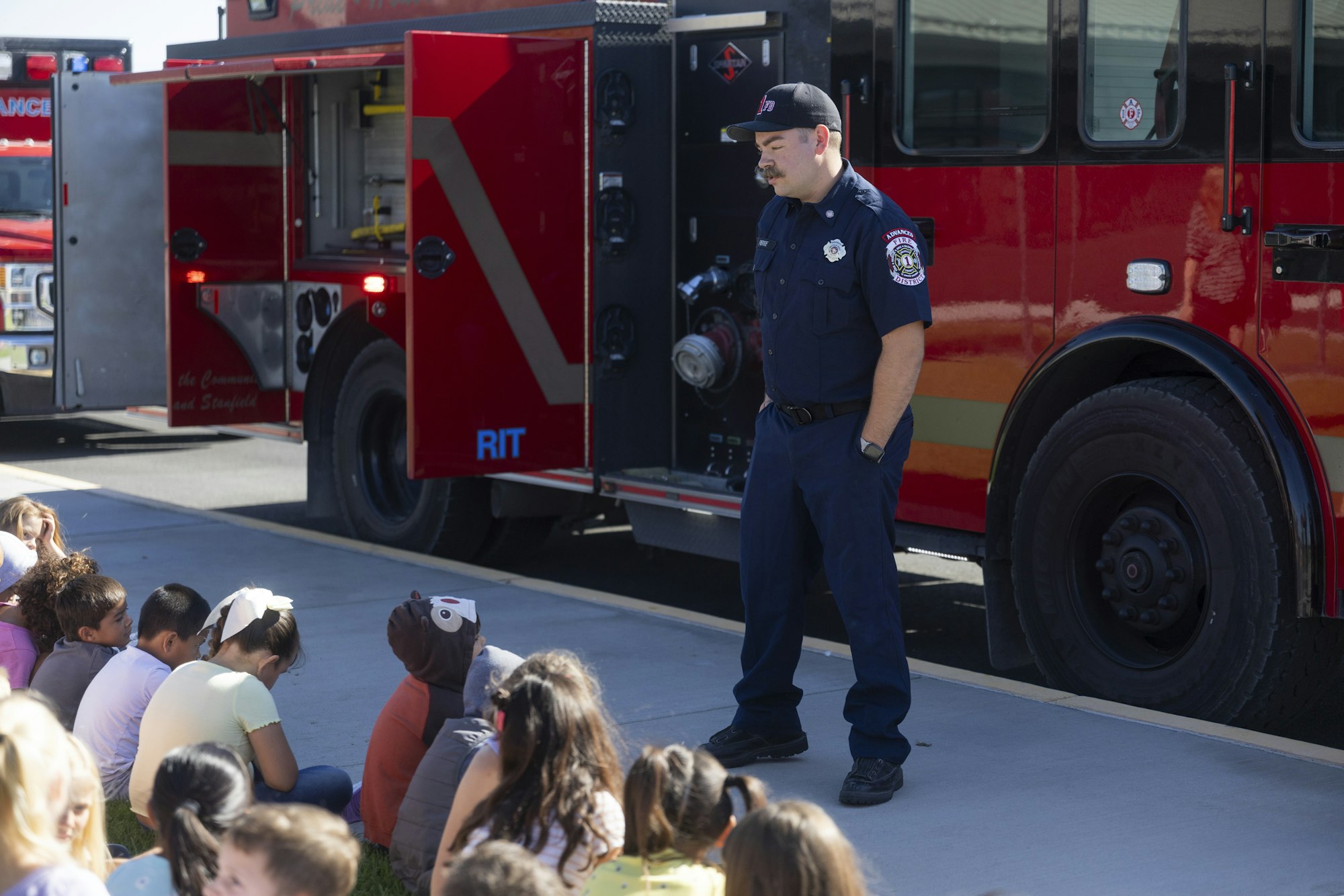 A firefighter stands beside a fire truck, engaging a group of seated children.