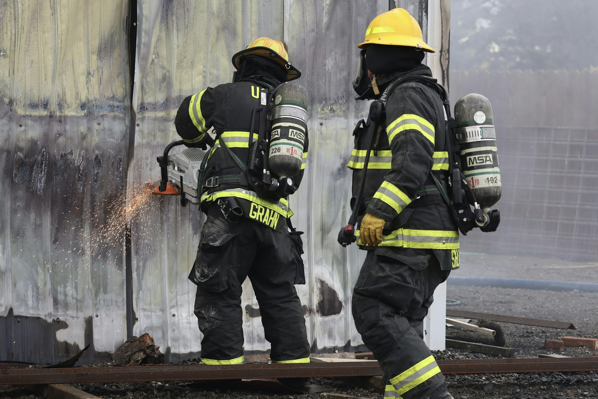 Firefighters in protective gear using a saw to cut through a metal wall, with sparks flying.