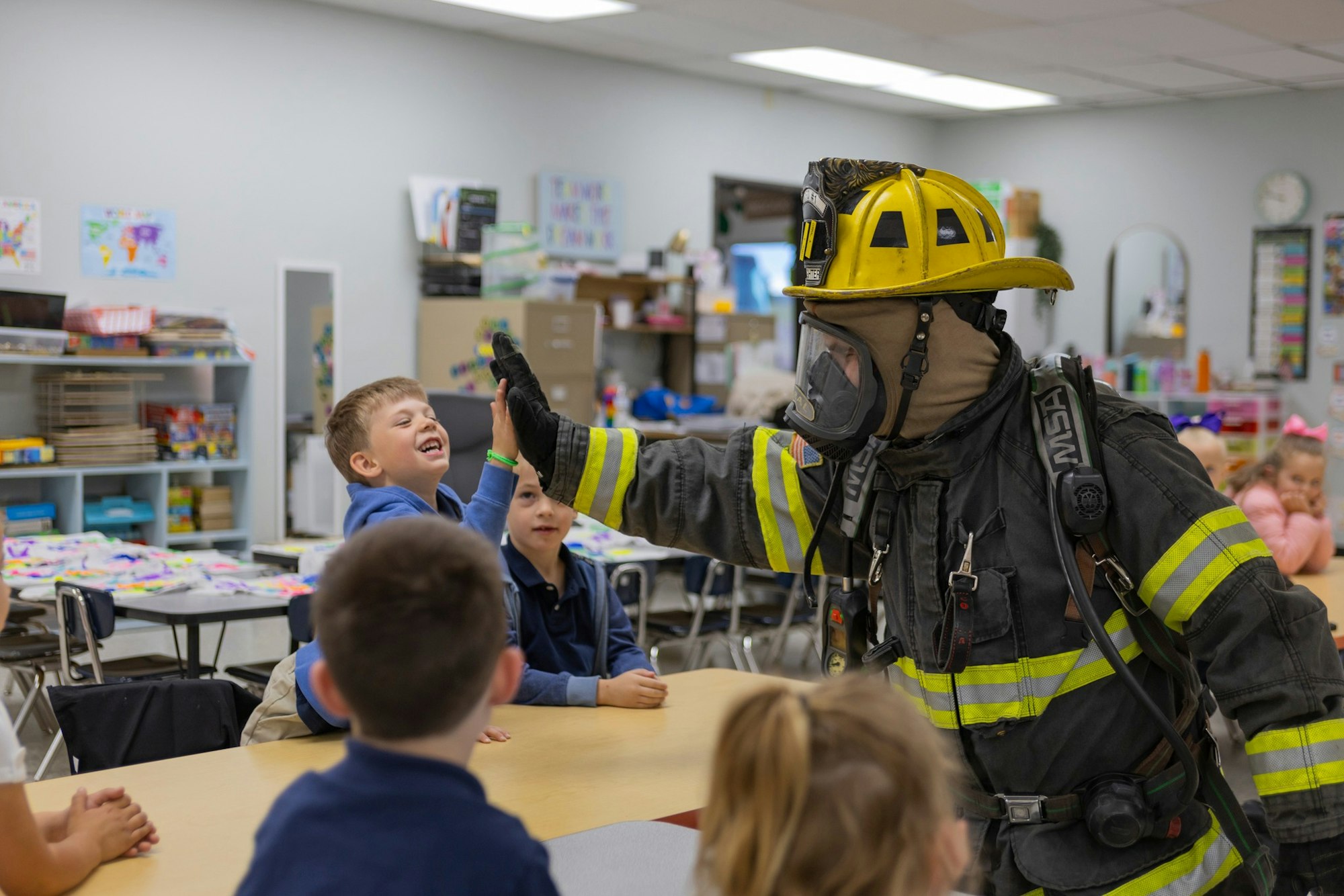 A firefighter gives a high-five to a laughing child in a classroom setting.