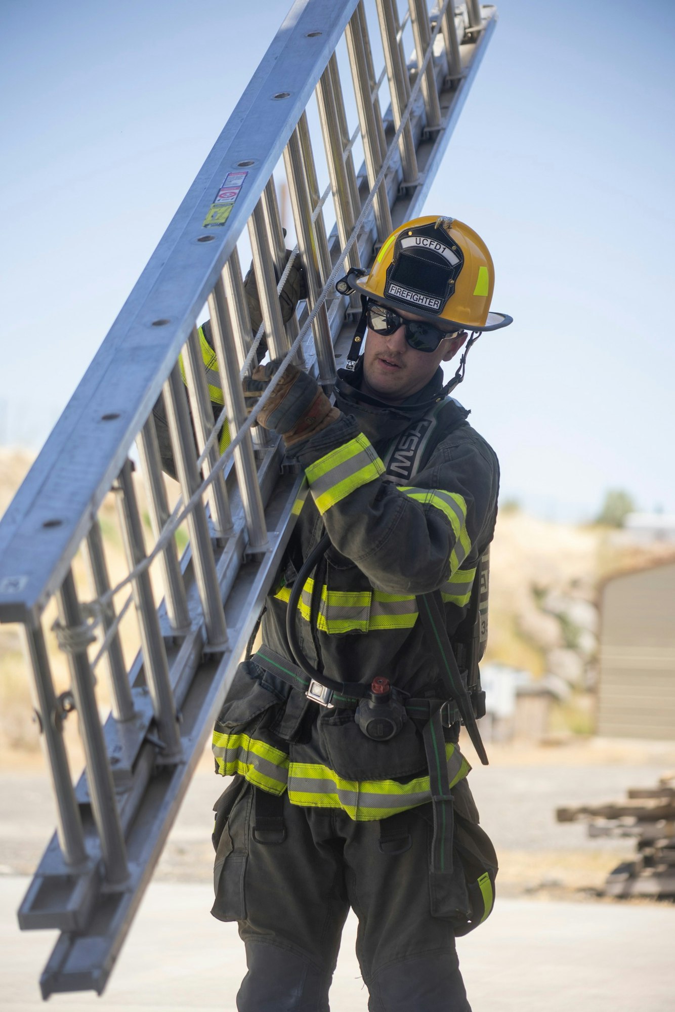 A firefighter wearing gear and sunglasses carries a metal ladder outdoors.