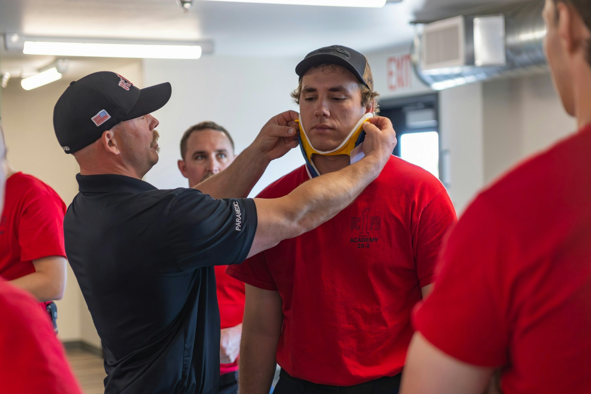 A paramedic helps a person with a cervical collar during training. Other people in red shirts are present.