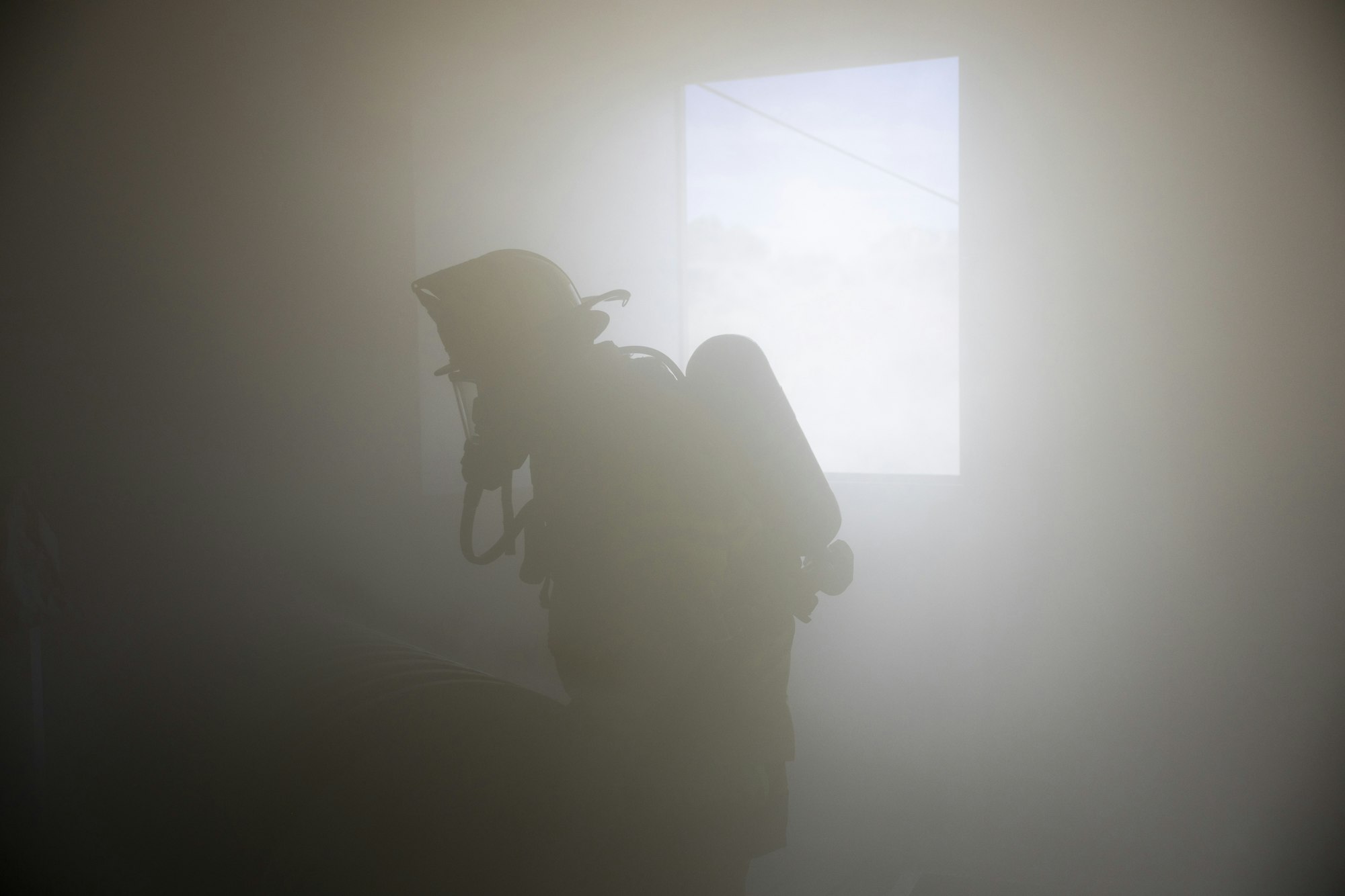 Silhouette of a firefighter with helmet and oxygen tank in a smoky environment near a bright window.