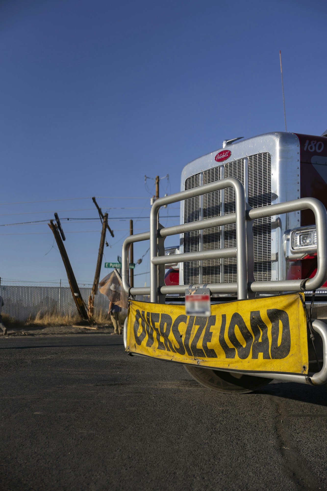 Truck with "OVERSIZE LOAD" sign, near tilted power pole under clear blue sky.