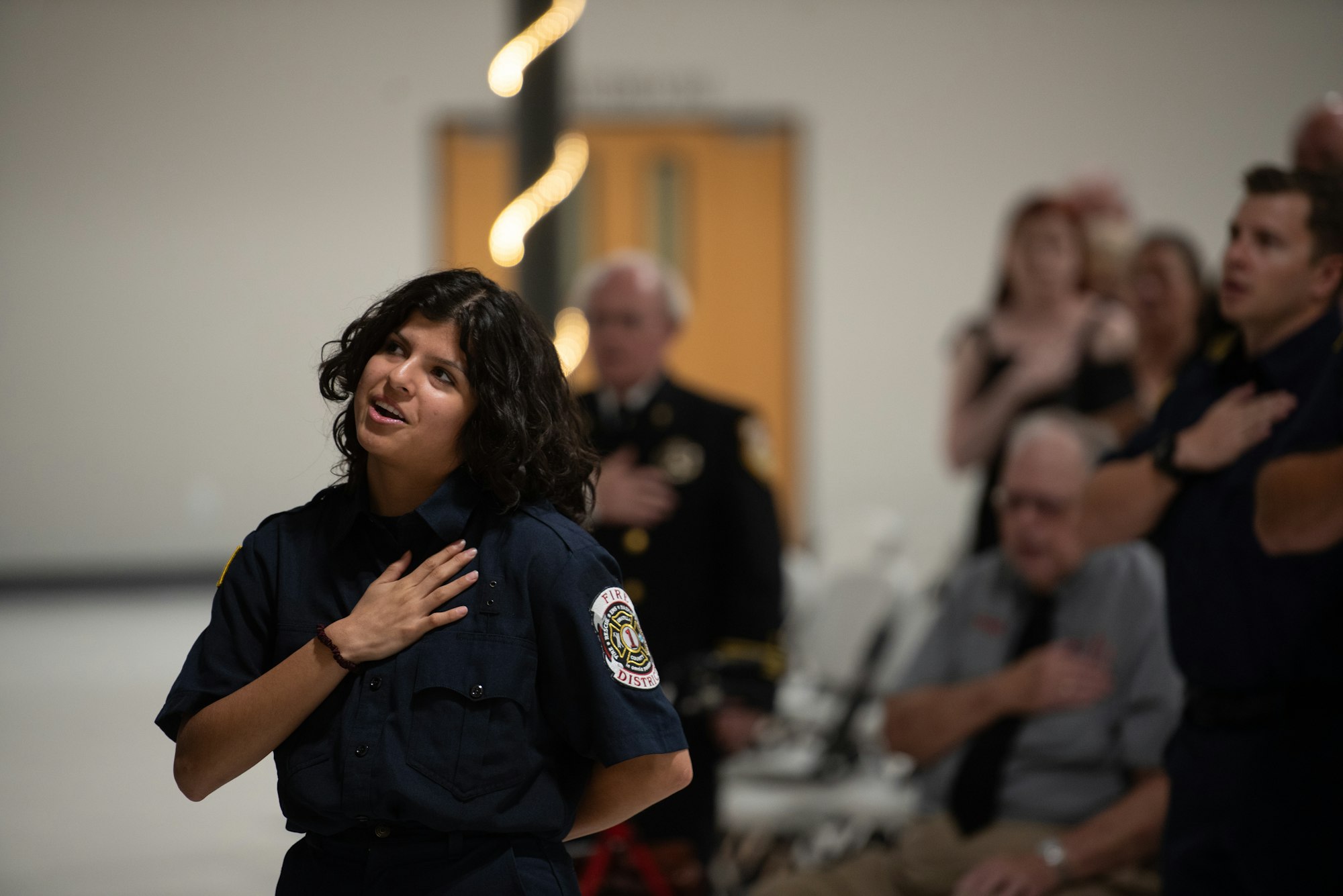 A person in a uniform stands with hand over heart, possibly during a pledge or anthem, with others in the background doing the same.
