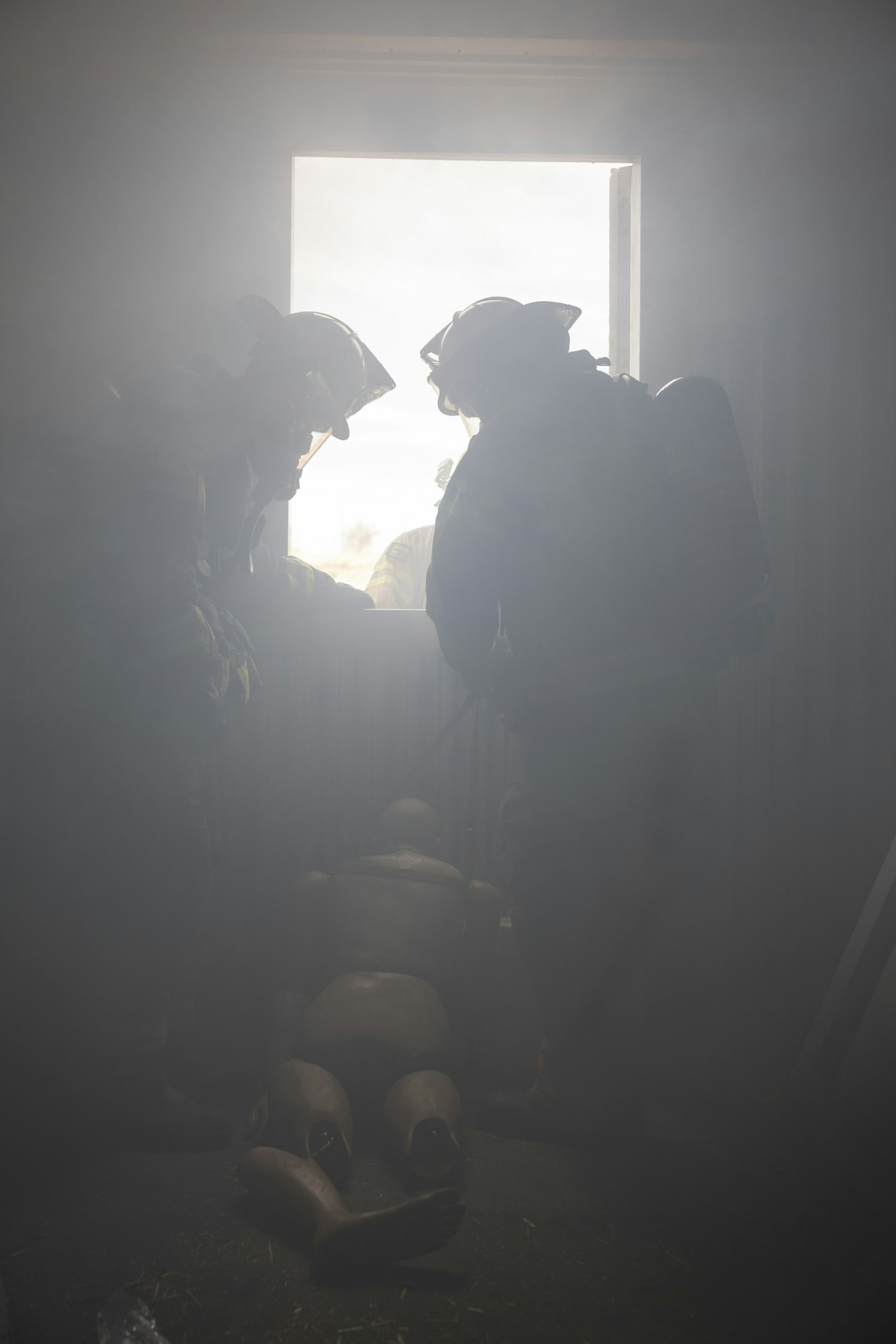 Two firefighters in gear are carrying a dummy through a smoky room towards a window.