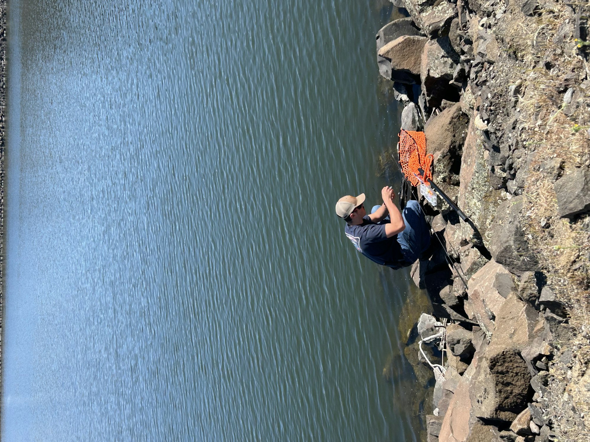 A person sitting on rocks by a body of water, holding a phone with an orange net and fishing gear nearby.