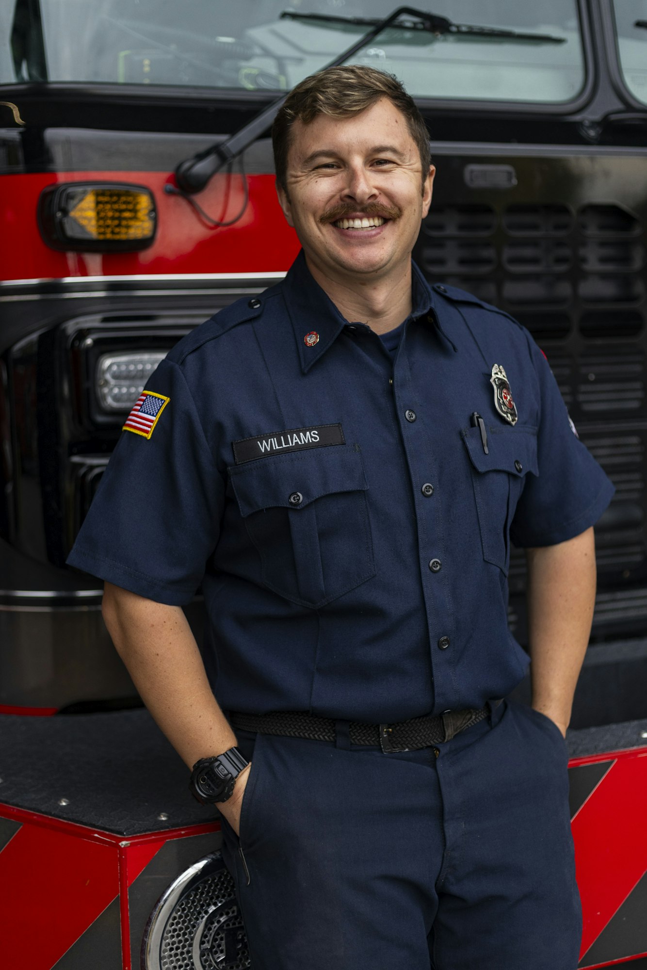 A firefighter in uniform stands in front of a fire truck, smiling.