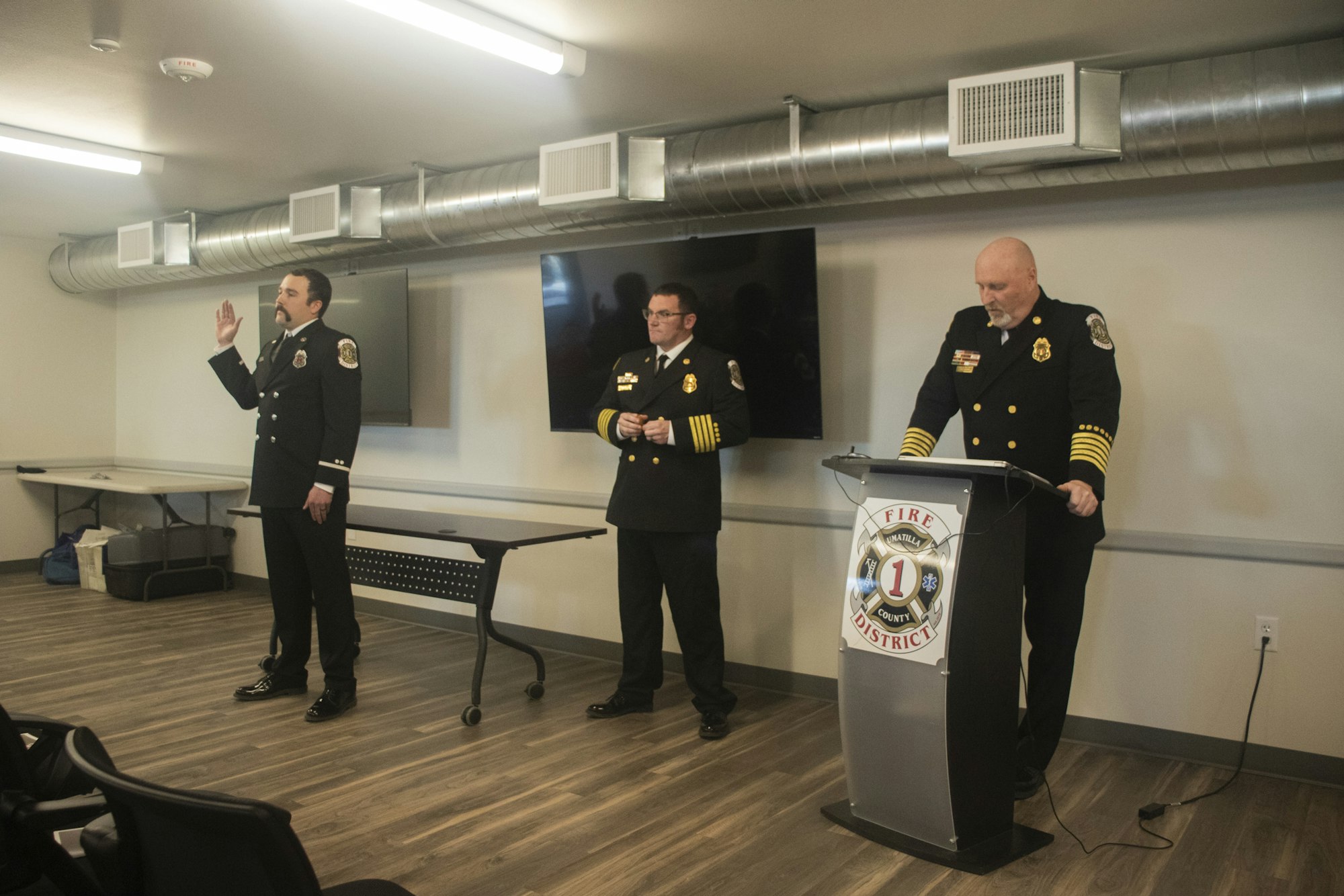 Three uniformed firefighters, one giving an oath, stand in a room with a podium and screen.