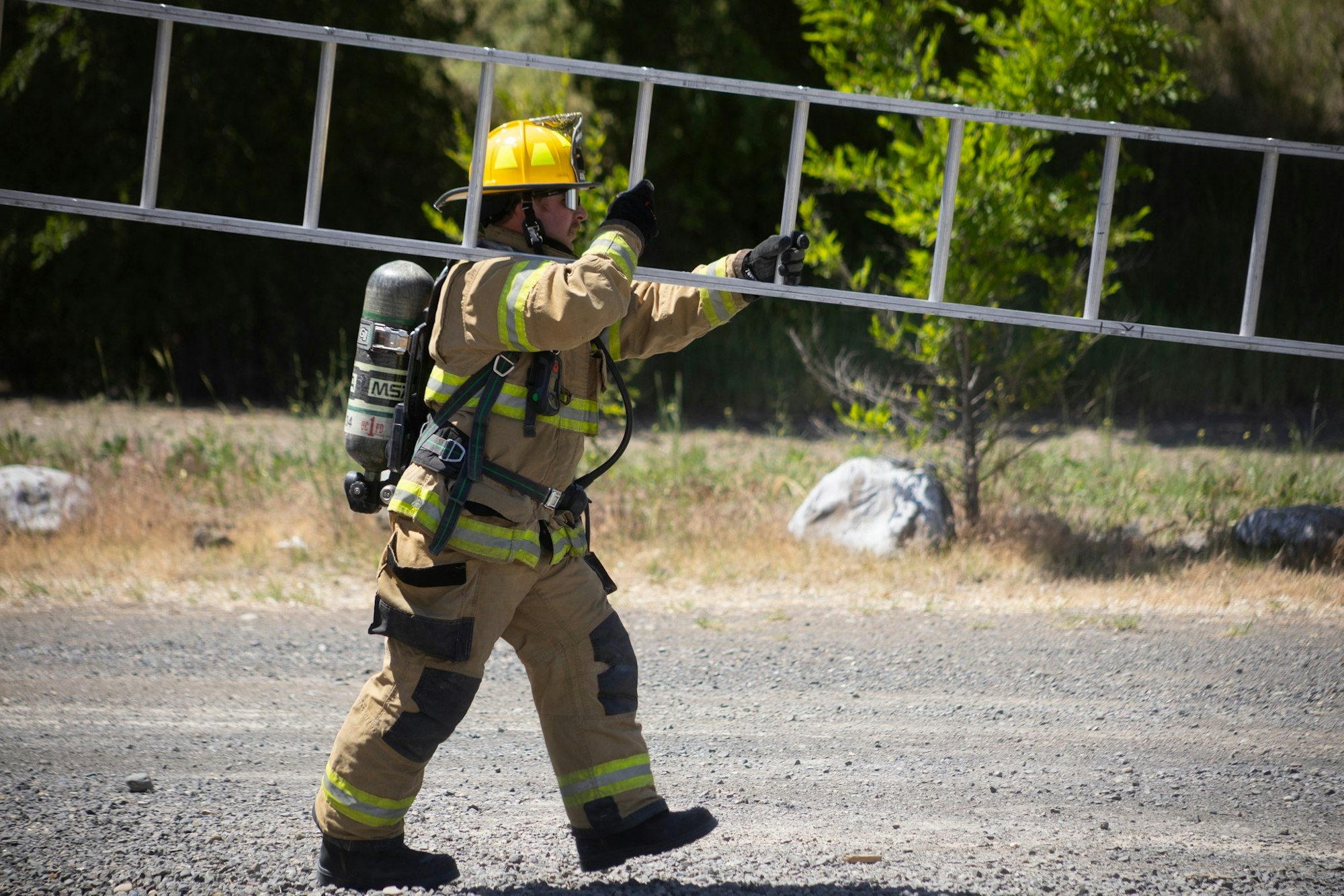 A firefighter in gear is carrying a ladder outdoors on a gravel surface.
