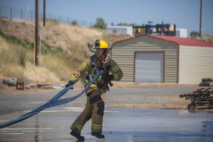 A firefighter in gear pulls a hose outdoors near a building.