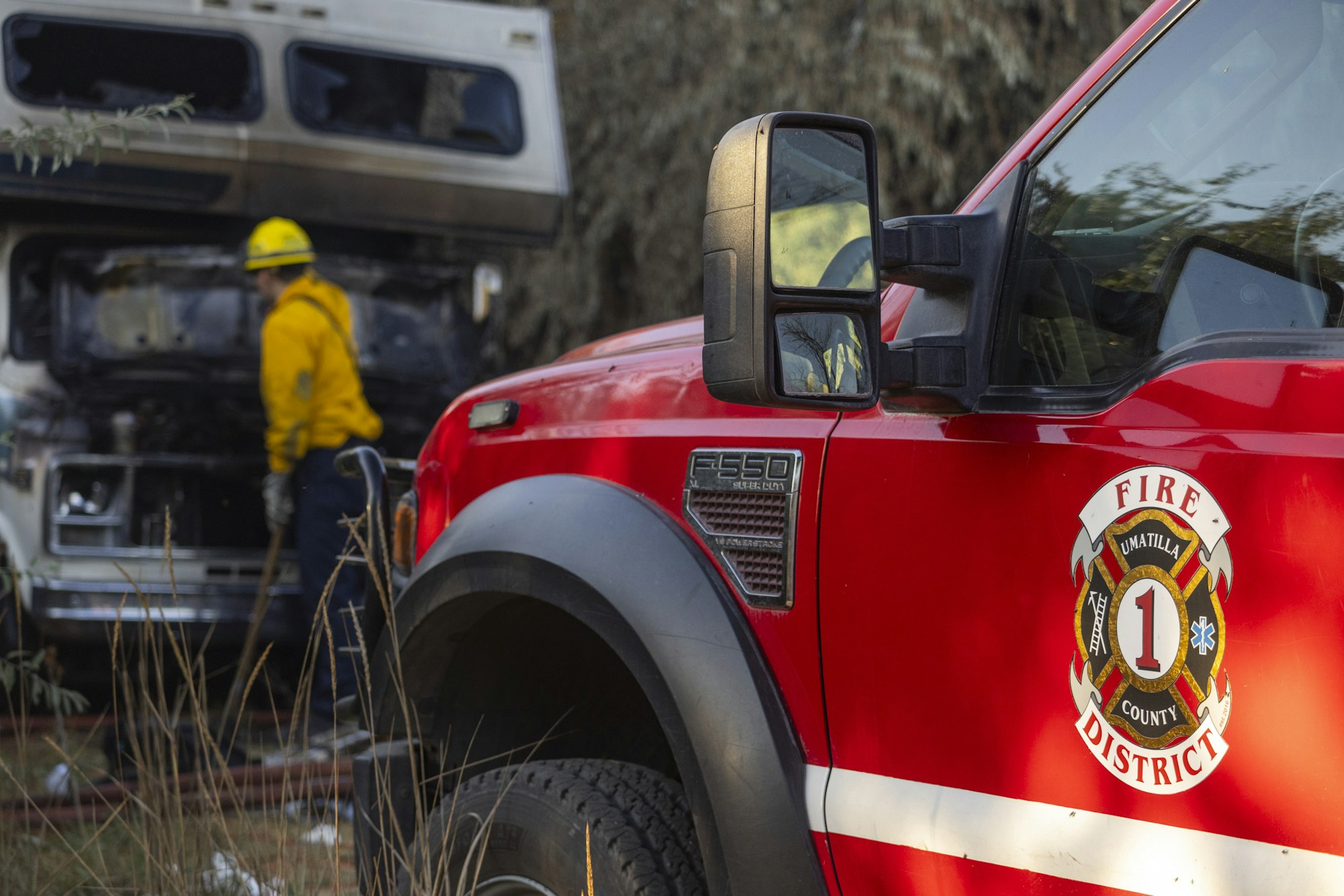 A firefighter near a red fire truck and a camper van, with trees in the background.