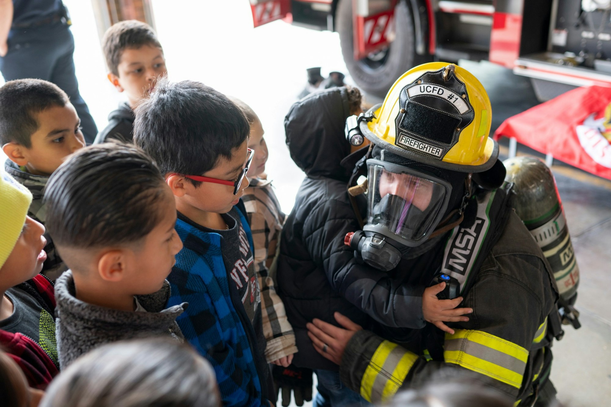 A firefighter in full gear interacts with a group of children, one hugging them, near a fire truck.