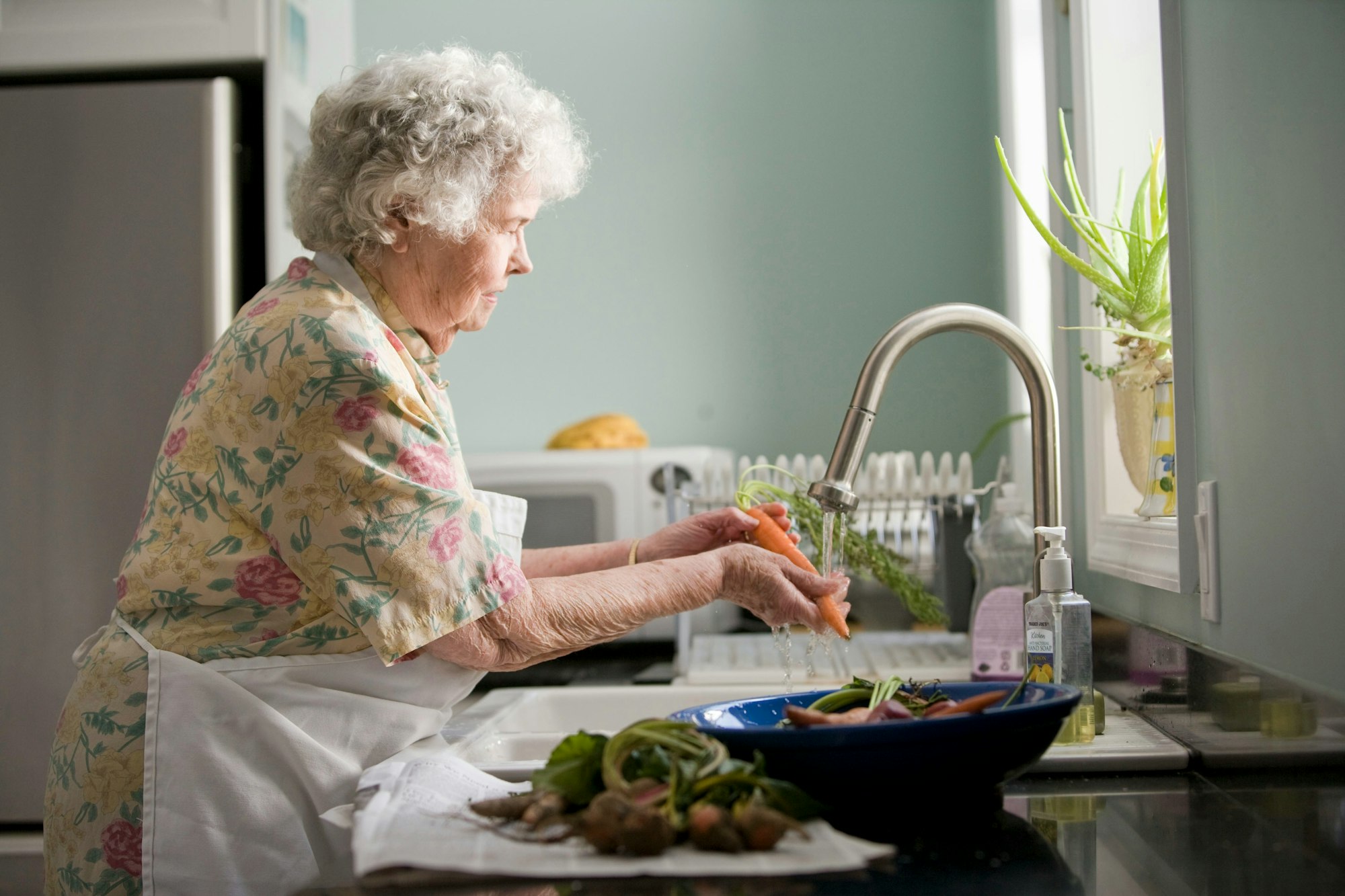 Elderly woman washing carrots at a kitchen sink.