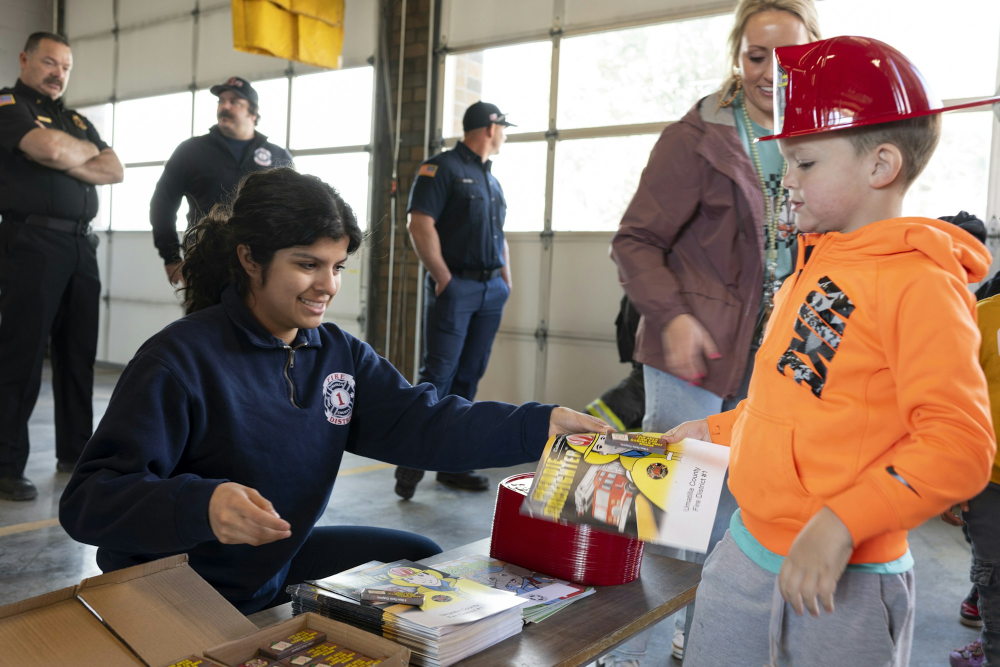A worker hands a child a booklet at an event, with other staff and attendees in the background.