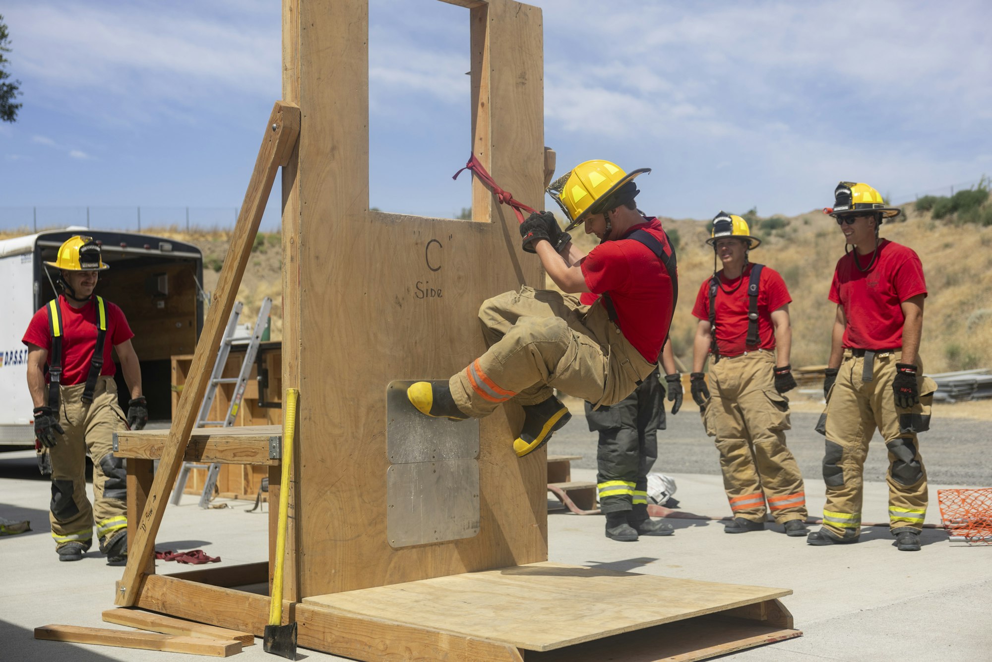 Firefighters training outdoor, one climbing through a wooden structure window, others watching.