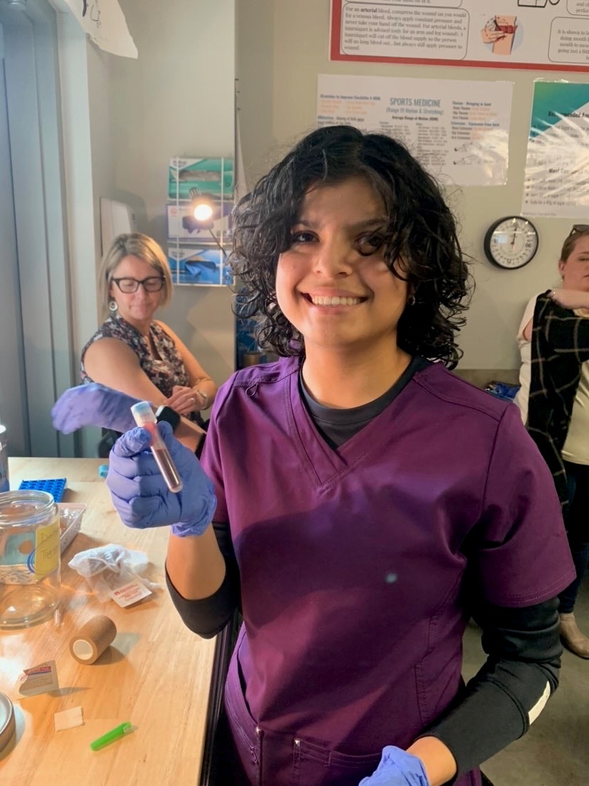 A person in purple scrubs smiling and holding a vial of blood in a lab setting.