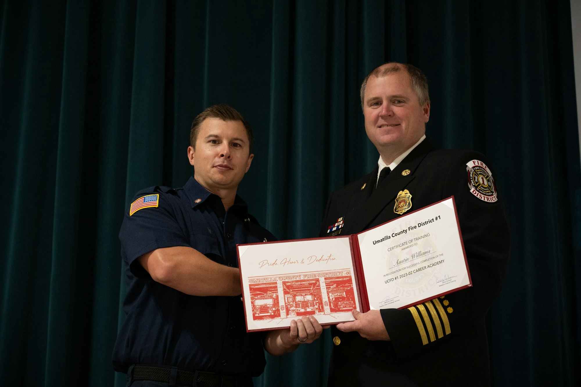 Two people in uniform in front of a curtain, holding a certificate from Umatilla County Fire District #1.