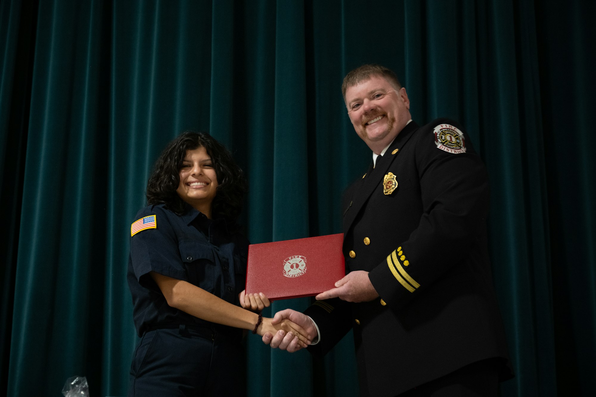 Two people shaking hands, smiling. One is in a uniform, holding a red diploma.