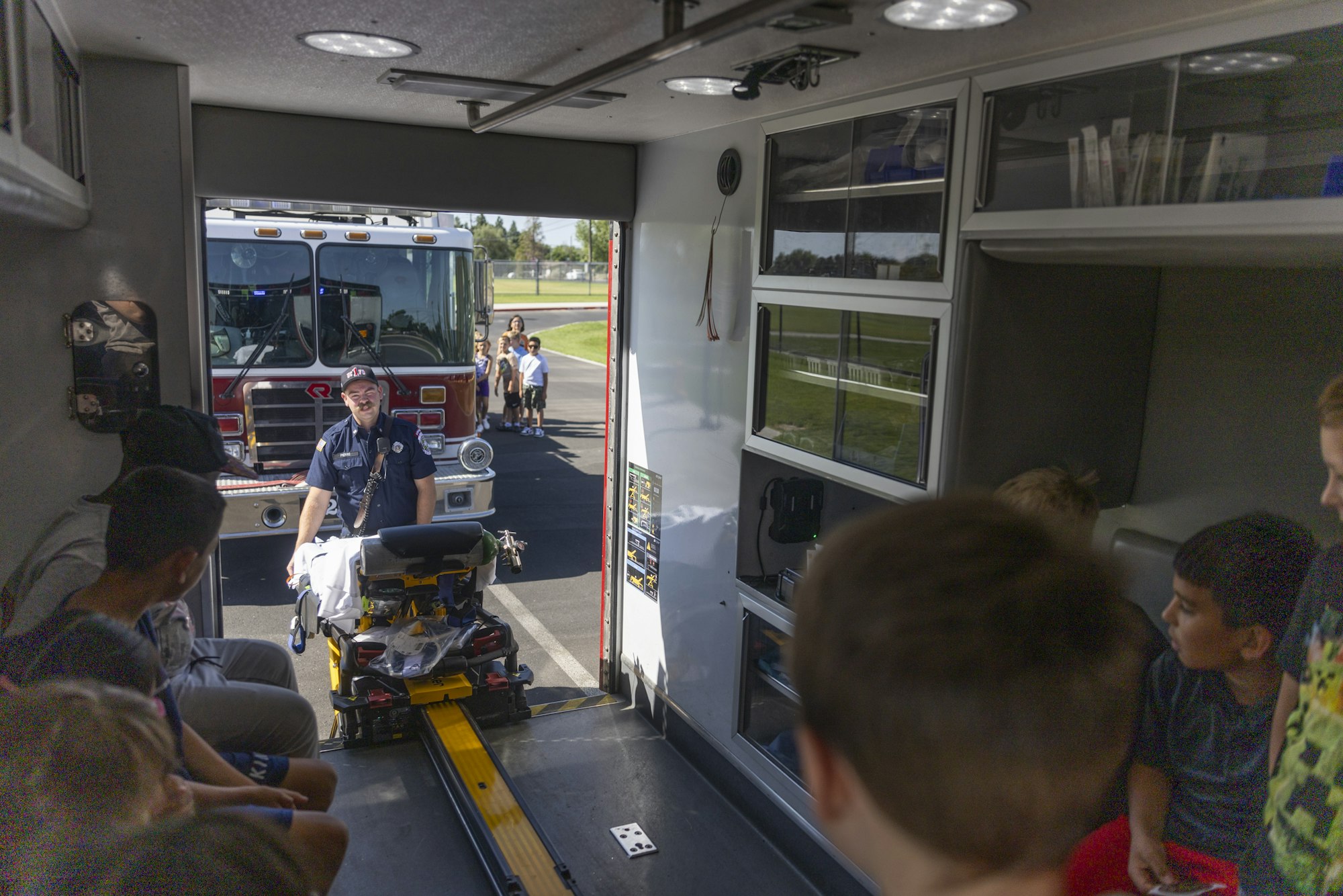 Children inside an ambulance, a firefighter with a stretcher outside, and a firetruck parked nearby.