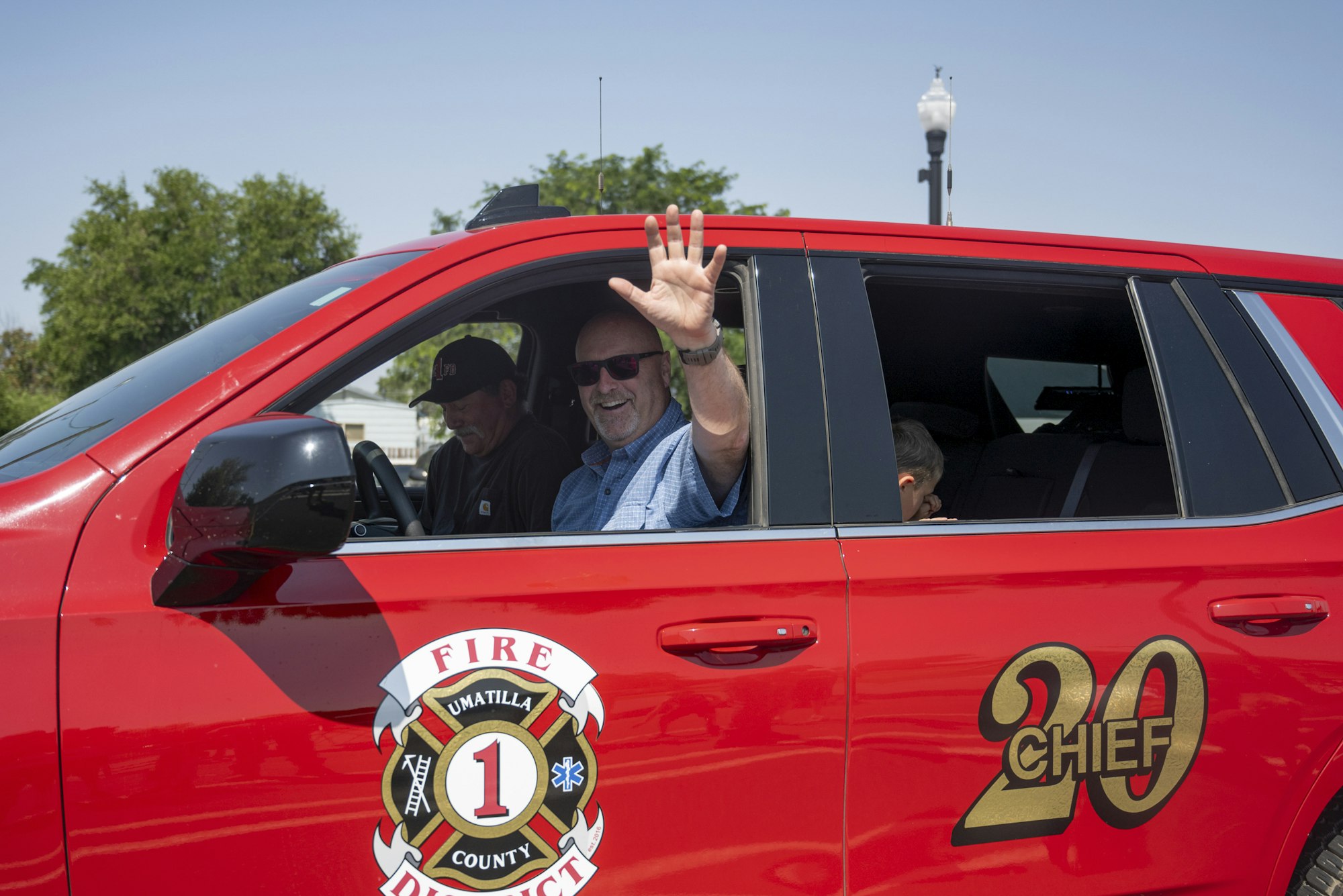 A person waves from a red Umatilla County fire chief vehicle, with two passengers inside.