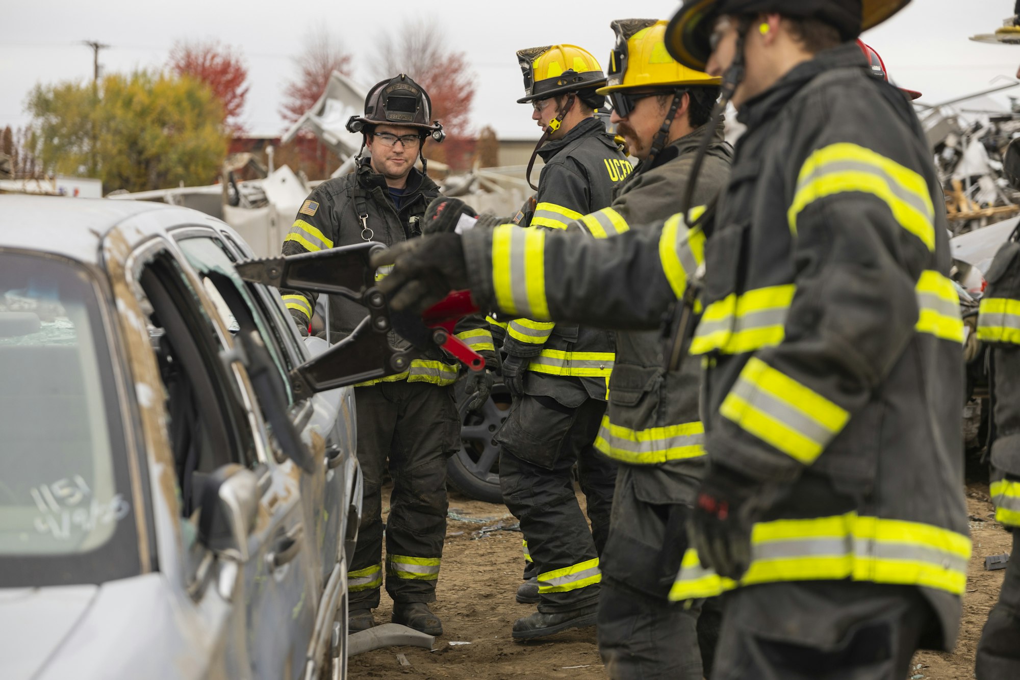 Firefighters in gear are using hydraulic tools on a car, likely practicing a rescue operation.