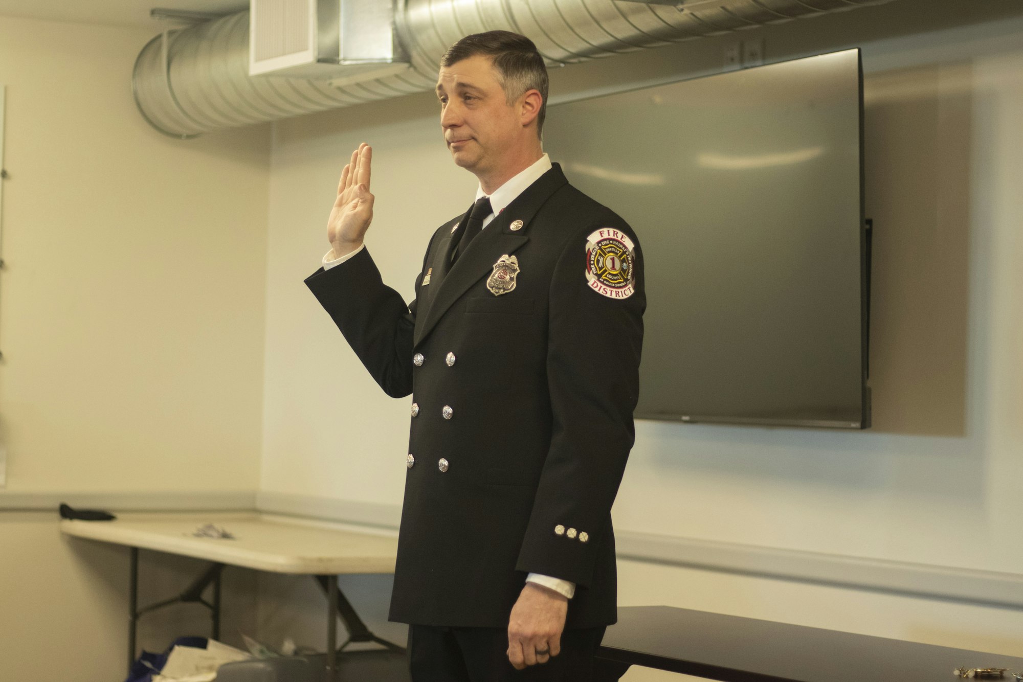 A firefighter in uniform is taking an oath with one hand raised, indoors.