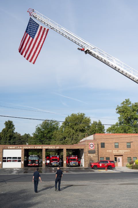 Fire station with fire trucks parked, American flag hanging from ladder, and two people outside.