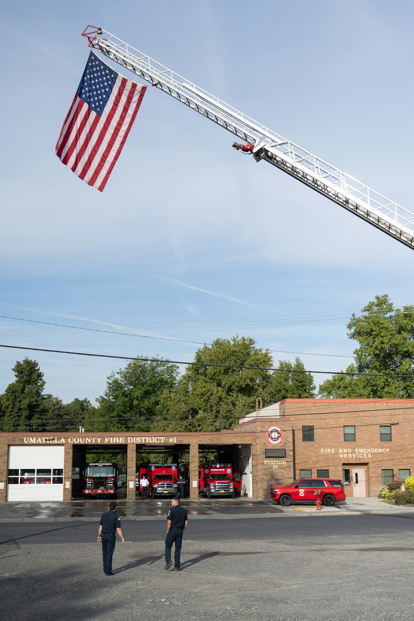 Fire station with fire trucks parked, American flag hanging from ladder, and two people outside.