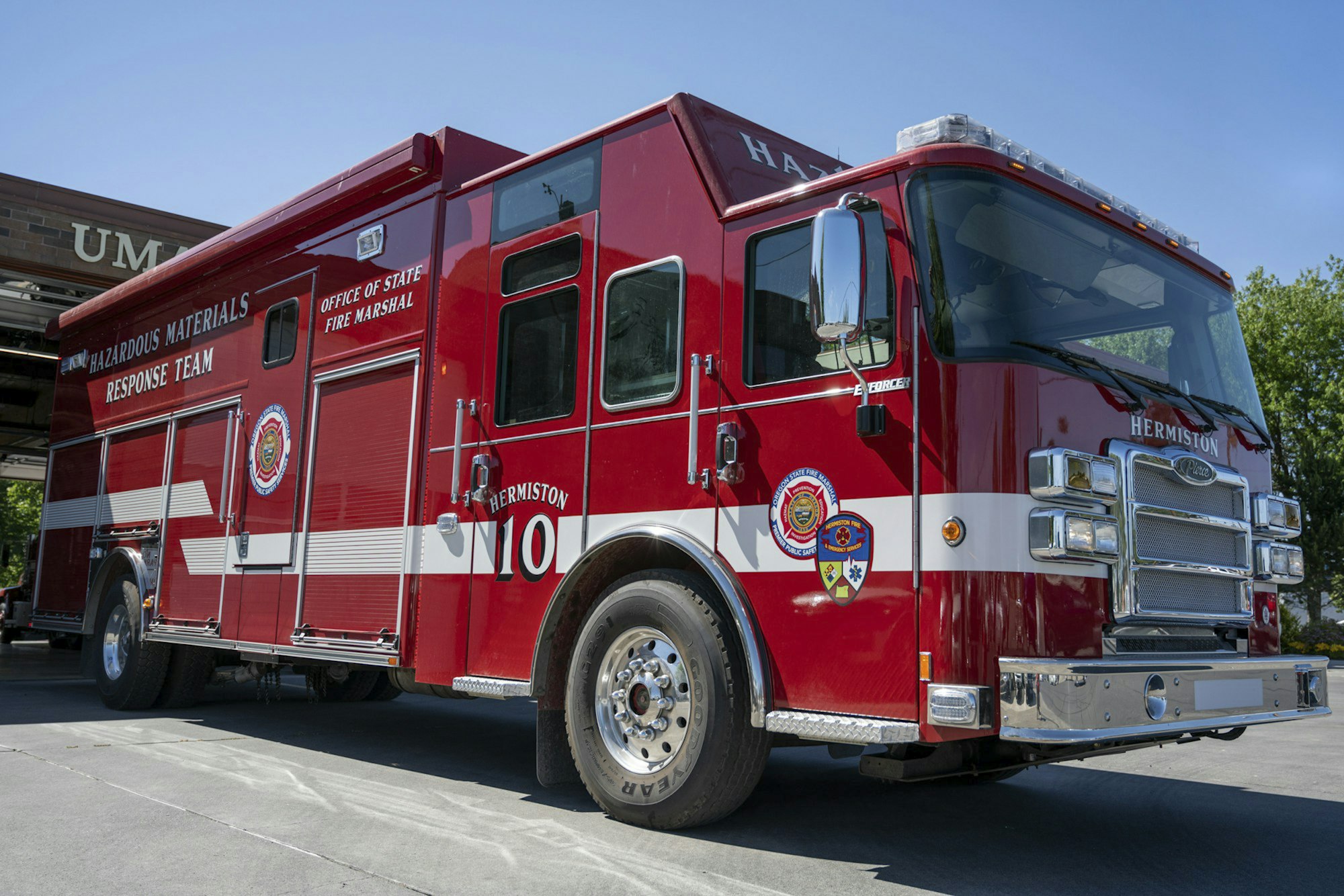 Red fire truck labeled "Hazardous Materials Response Team" with various department logos, parked outside a garage.